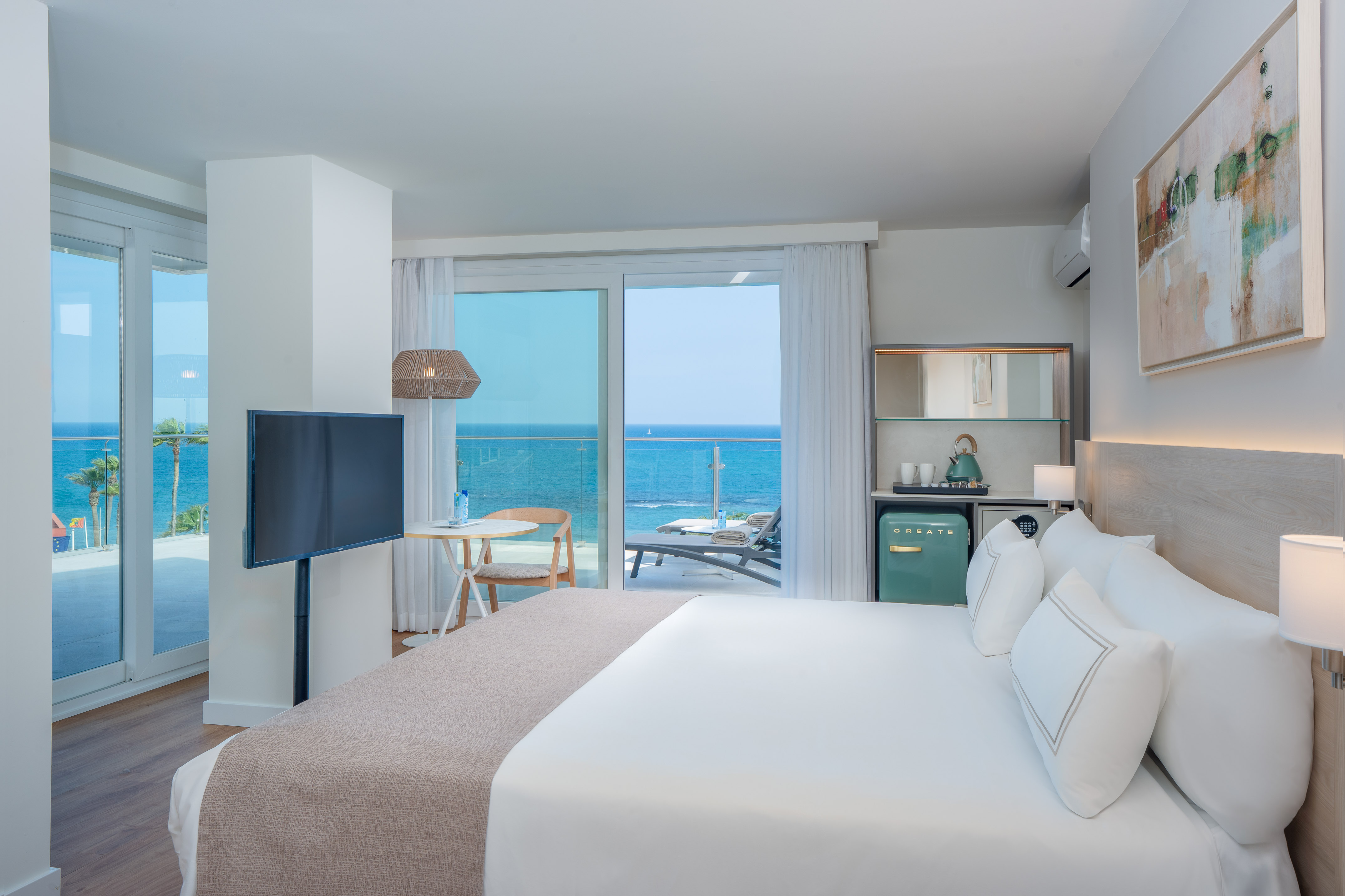Modern hotel room with ocean view balcony, large bed, TV, and mini-bar.