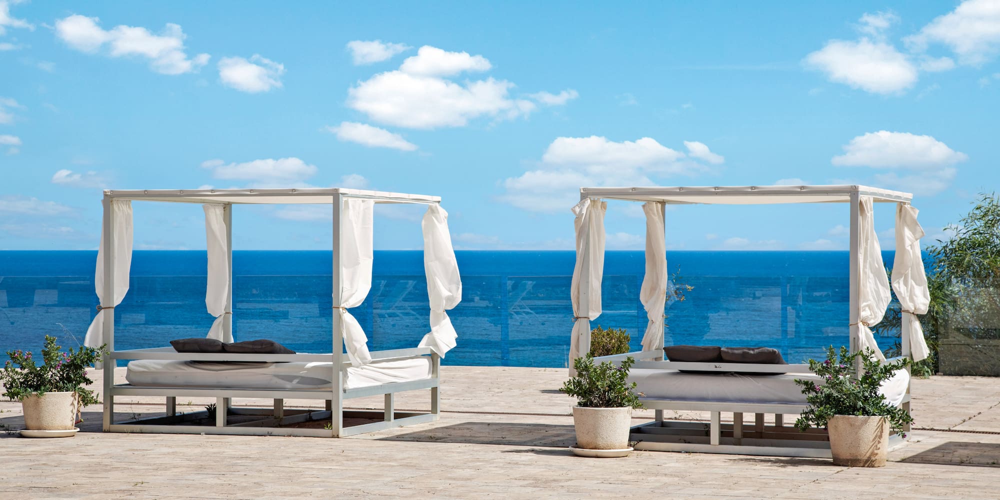 Two white canopy daybeds on an outdoor patio overlooking the blue ocean.