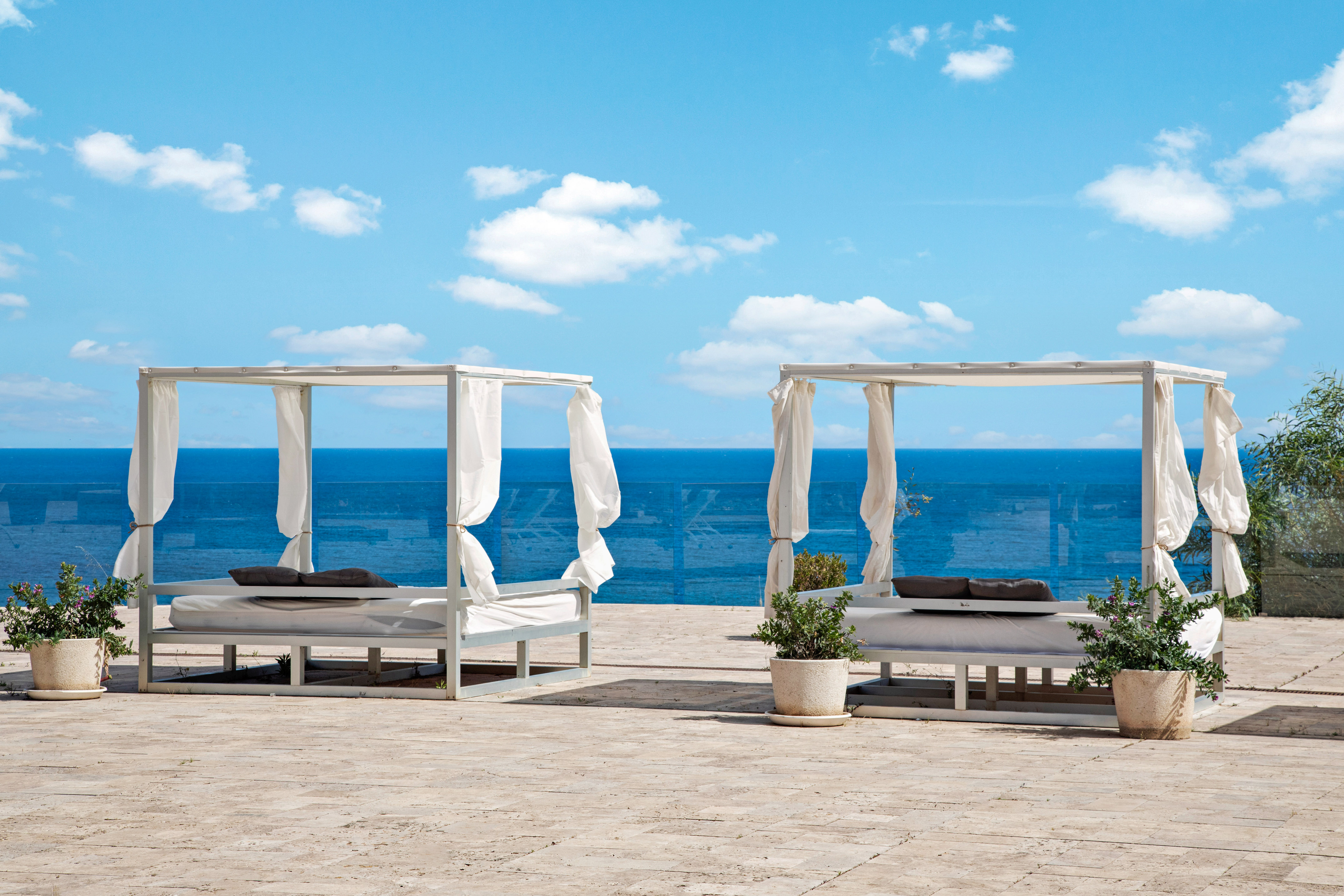 Two white canopy daybeds on an outdoor patio overlooking the blue ocean.