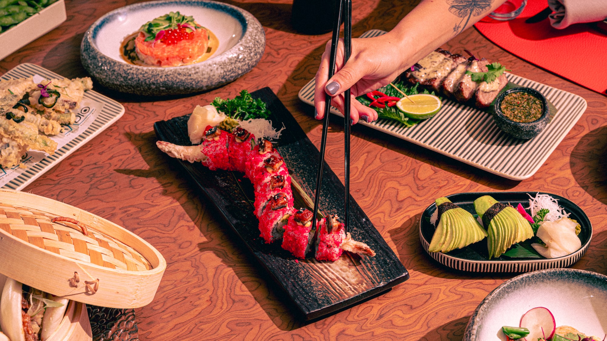 Hand picking a red sushi roll with chopsticks from a diverse Japanese food spread.