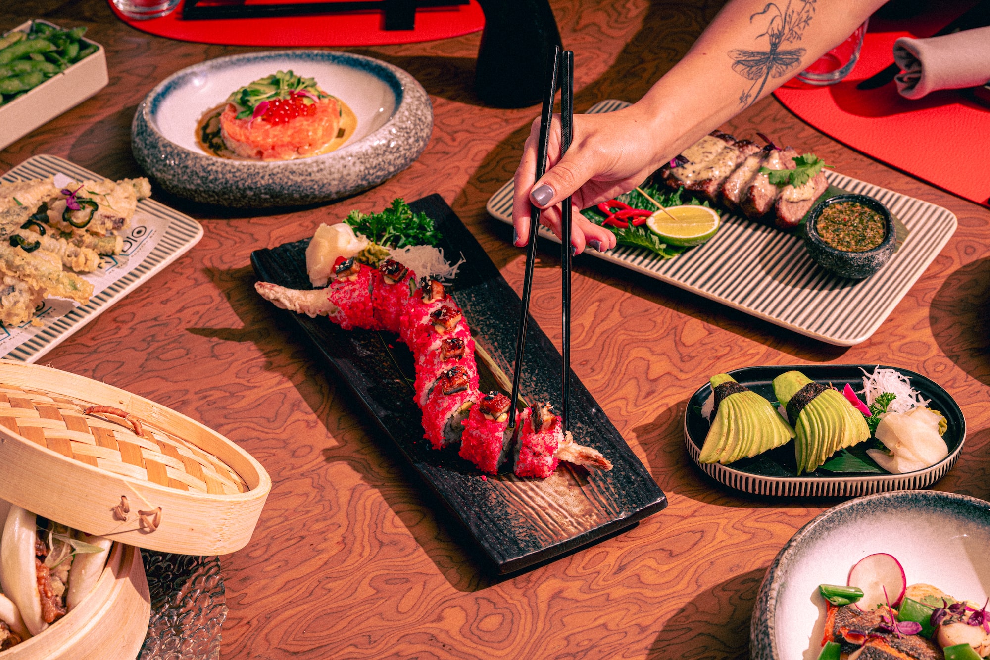 Hand picking a red sushi roll with chopsticks from a diverse Japanese food spread.