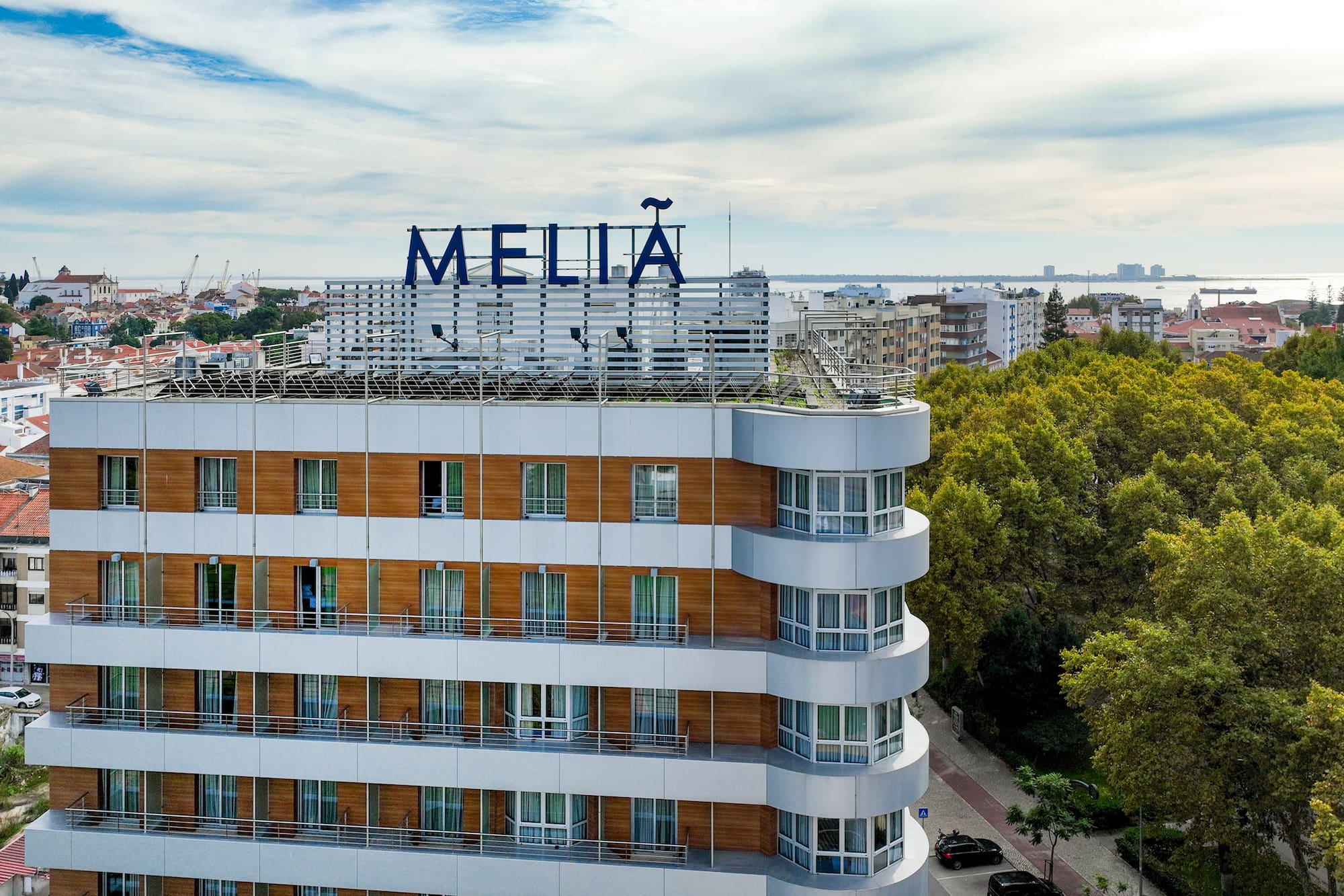 Melia hotel, modern facade, balconies, cityscape with green trees and waterfront.