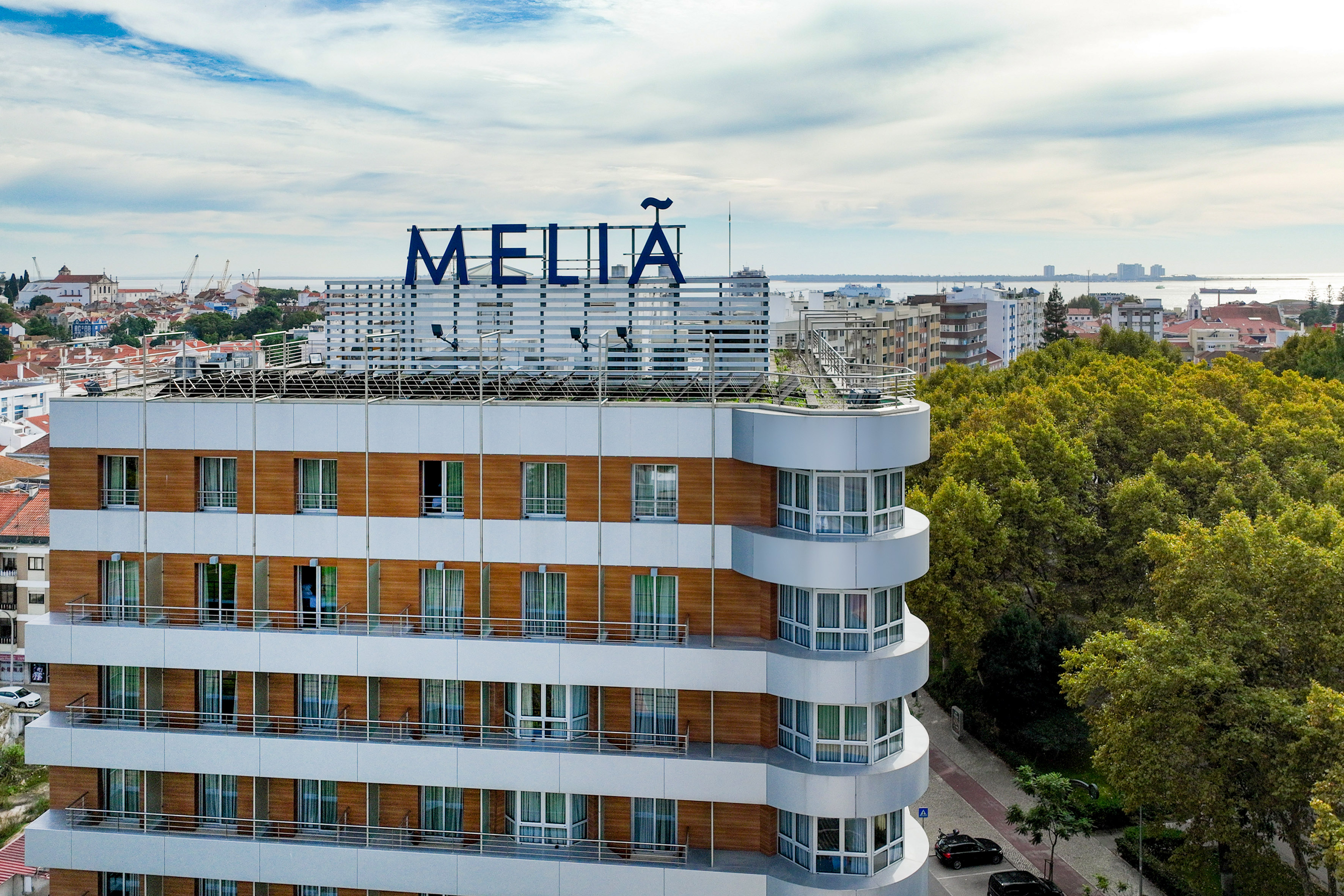 Melia hotel, modern facade, balconies, cityscape with green trees and waterfront.