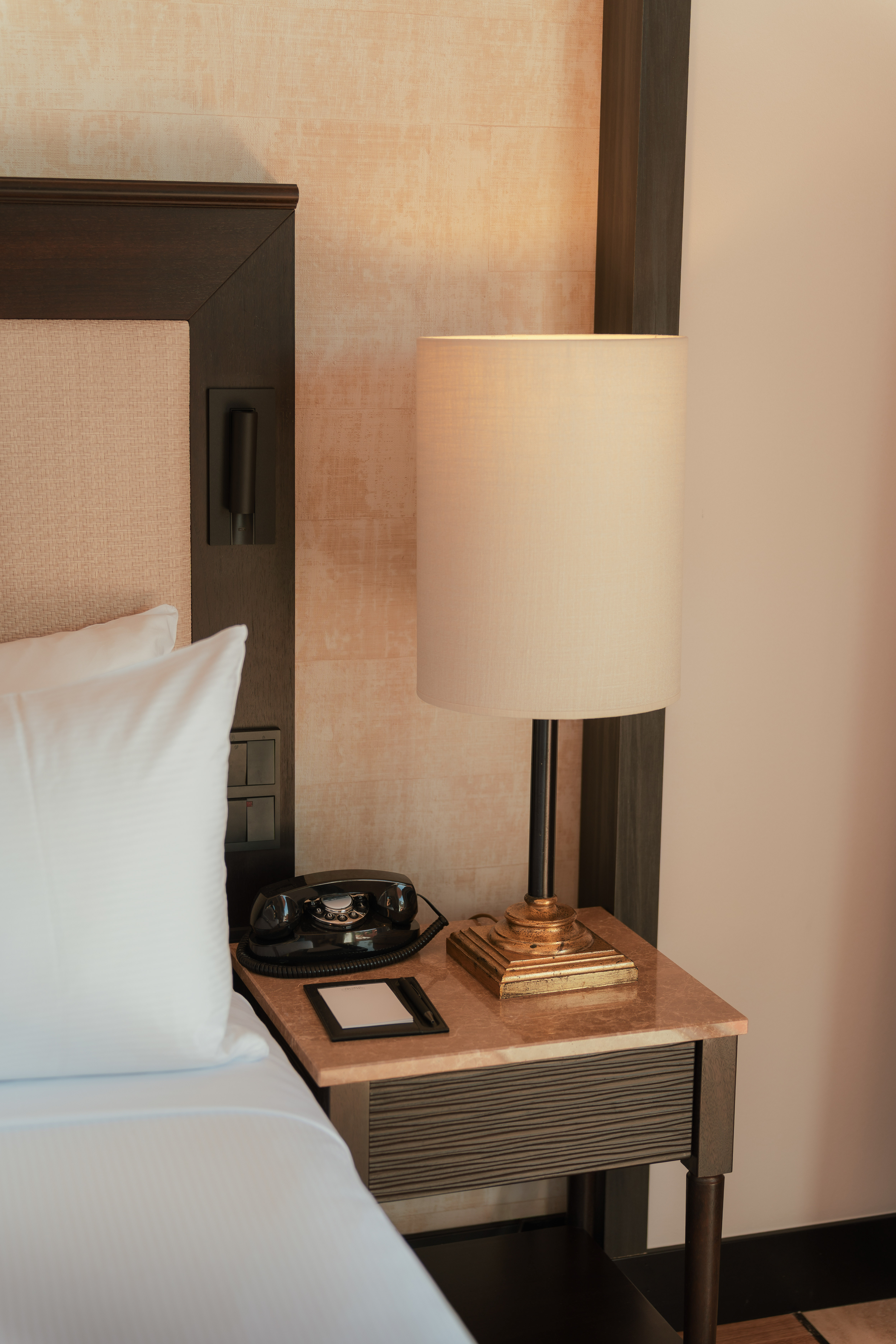 Hotel room bedside table with vintage rotary phone, illuminated lamp, notepad.