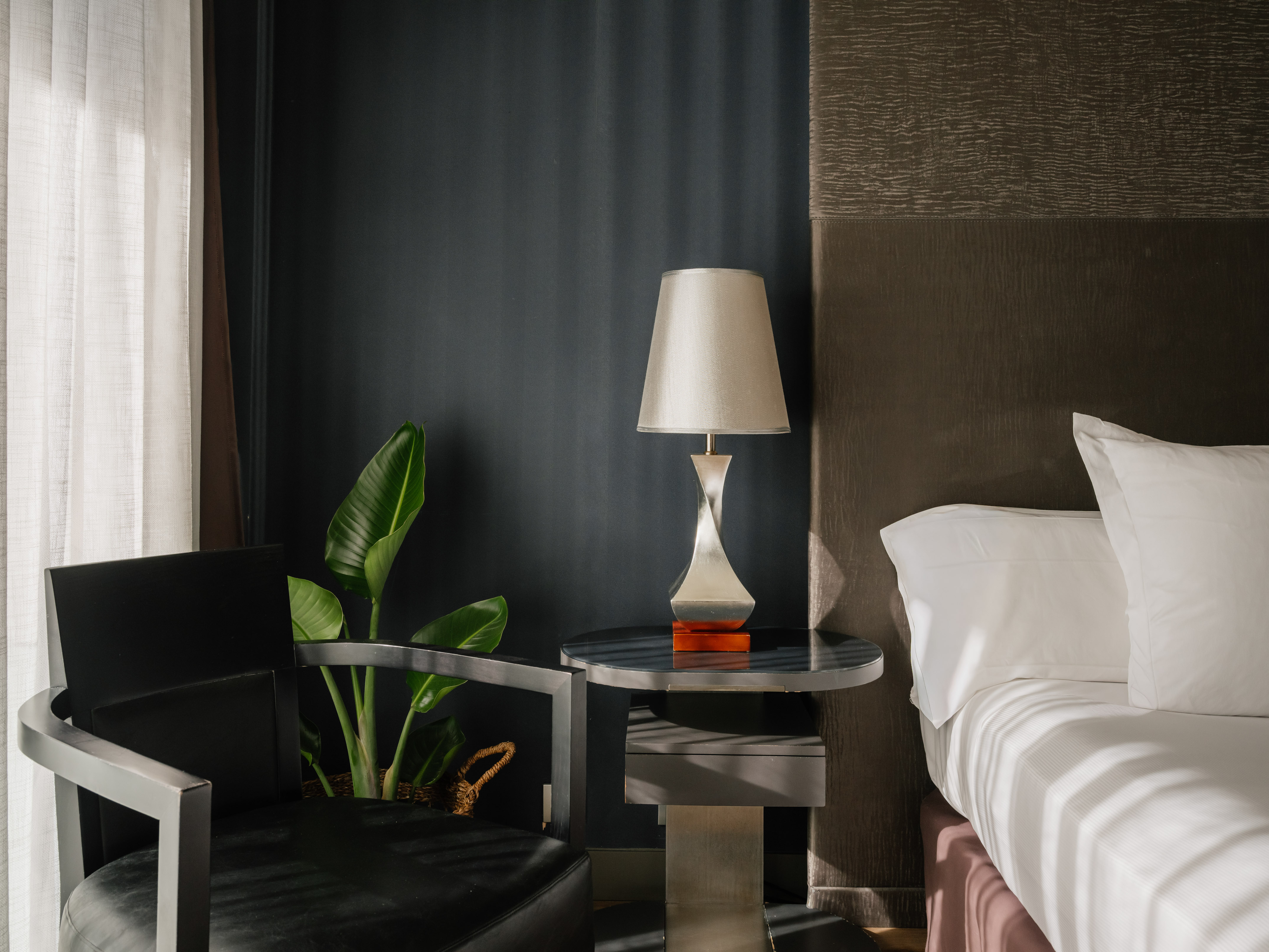 Dark modern bedroom with bed, bedside lamp, accent chair, and plant.