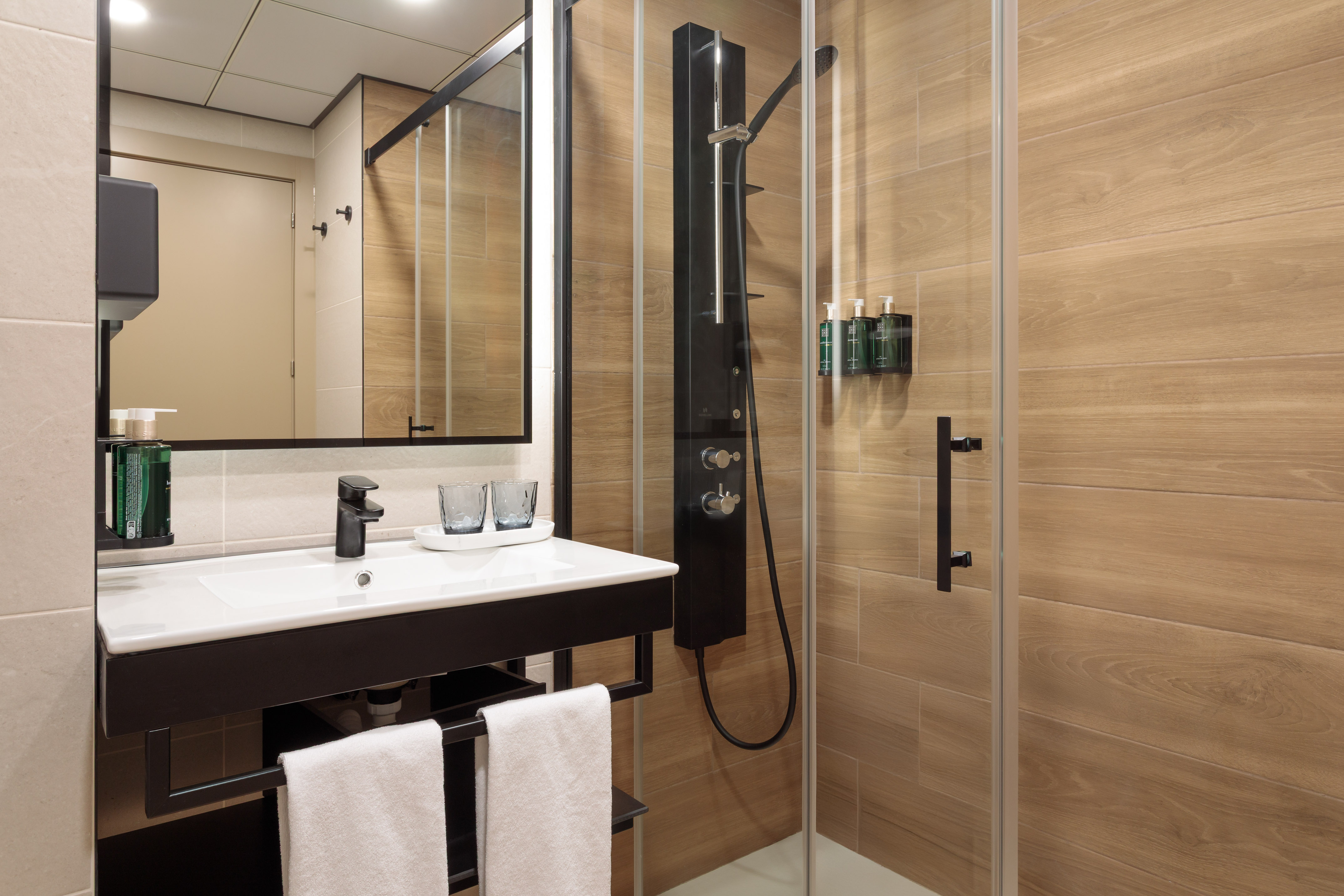 Modern hotel bathroom with black vanity, white sink, and glass shower, wood tiles.