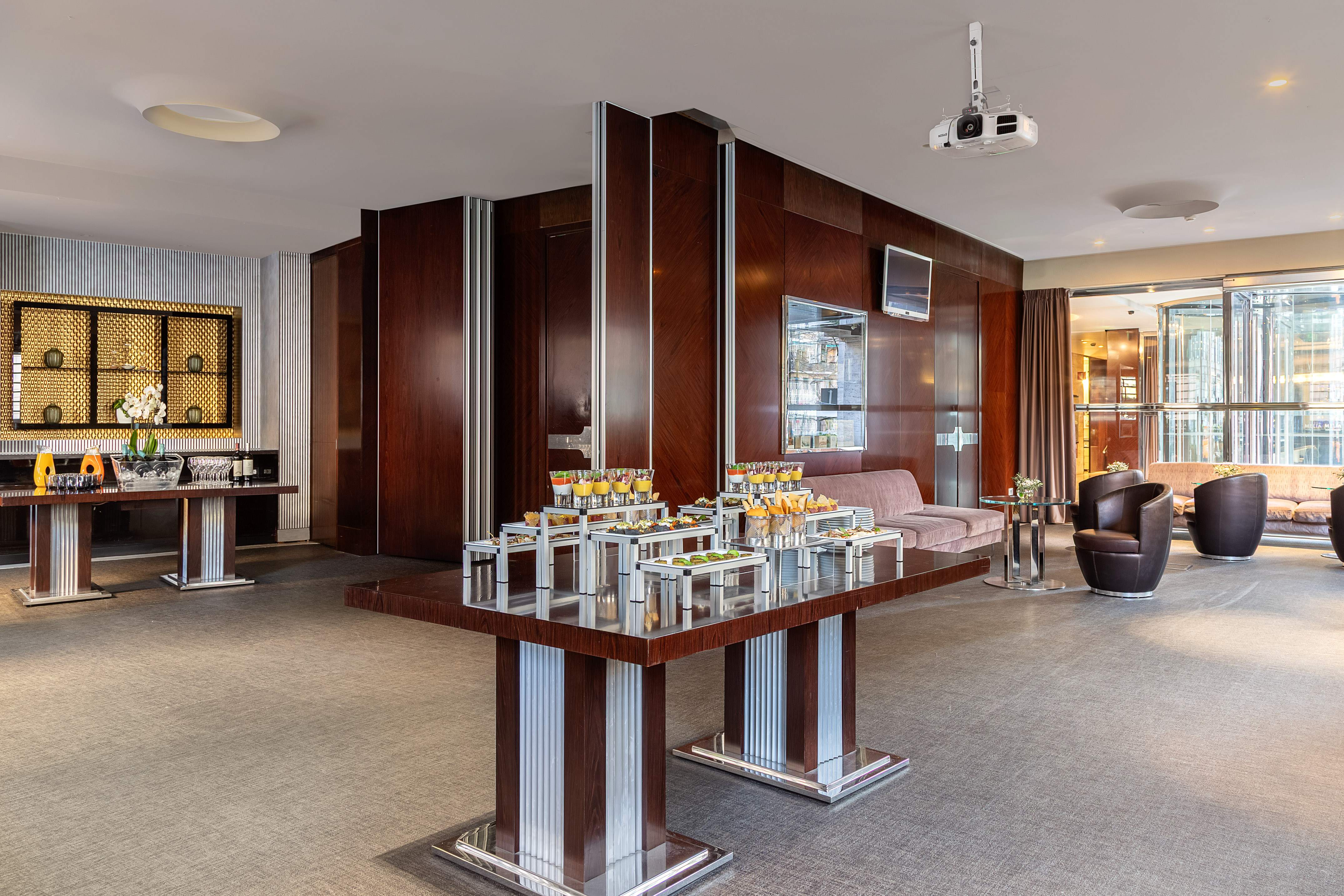 Modern hotel lounge with buffet, dark wood paneling, projector, and seating areas.