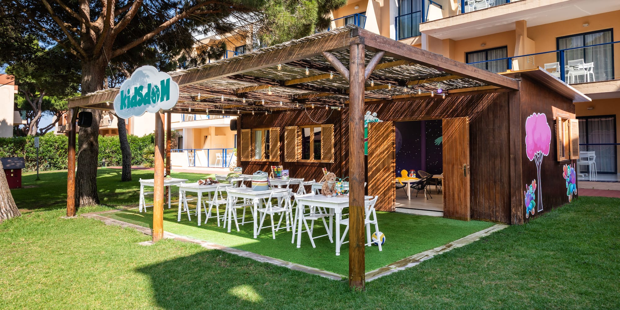 Resort kids club outdoor play area with wooden hut, tables, and KidsDom sign.