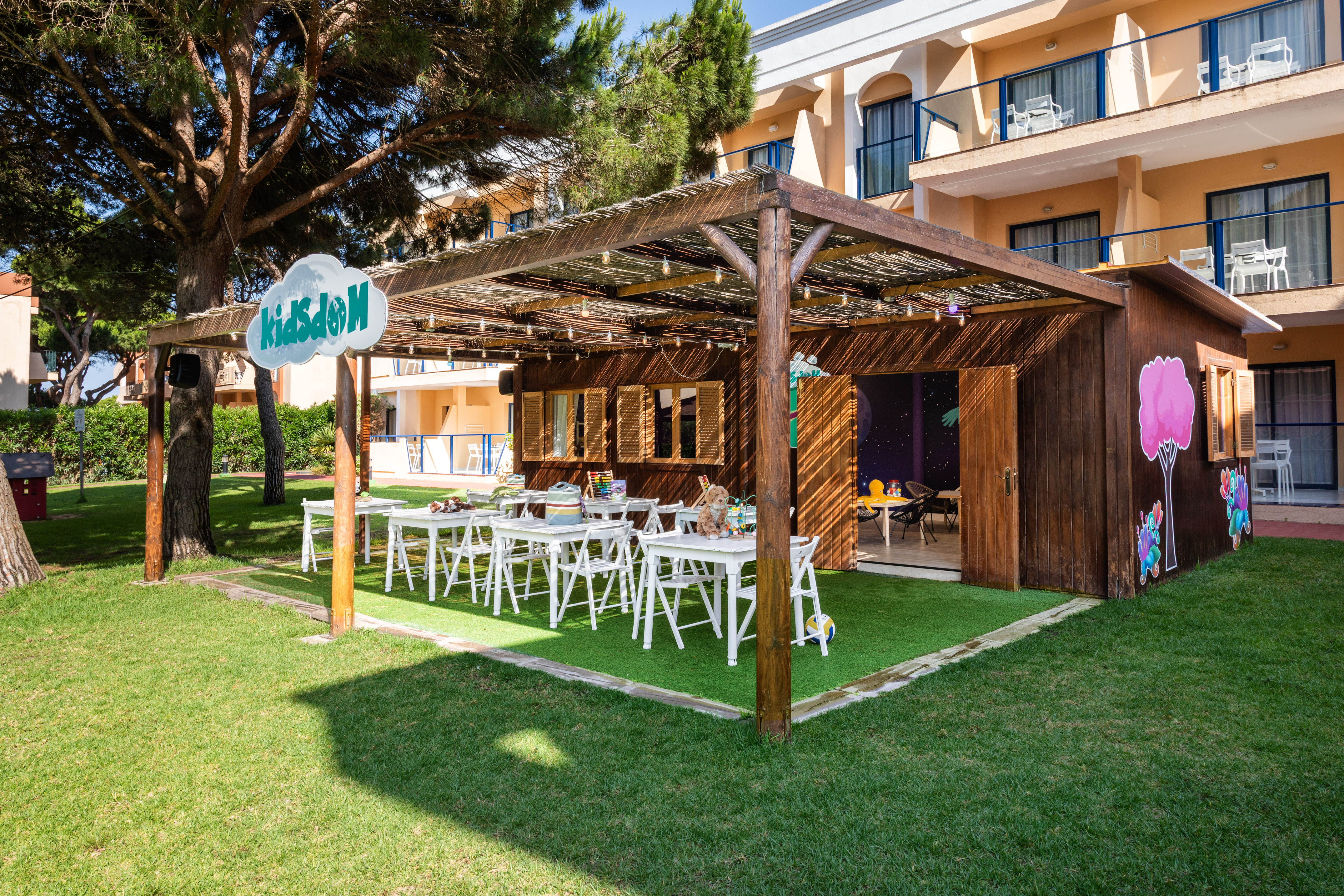 KidsDom outdoor club with wooden shelter, activity tables, and green lawn.