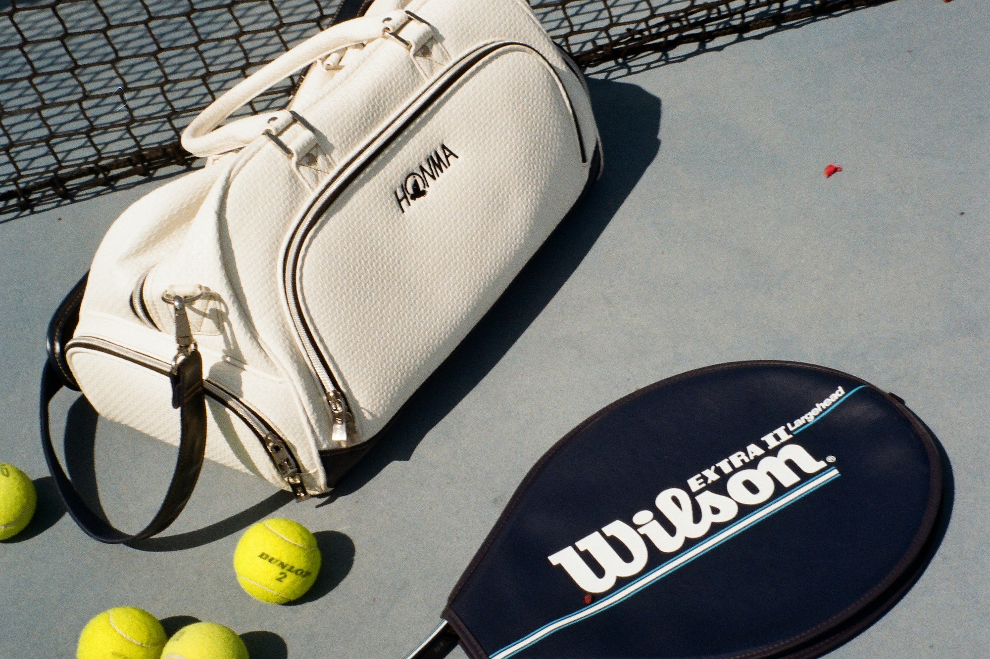 Equipo de tenis: bolsa Honma blanca, raqueta Wilson azul y pelotas en cancha.