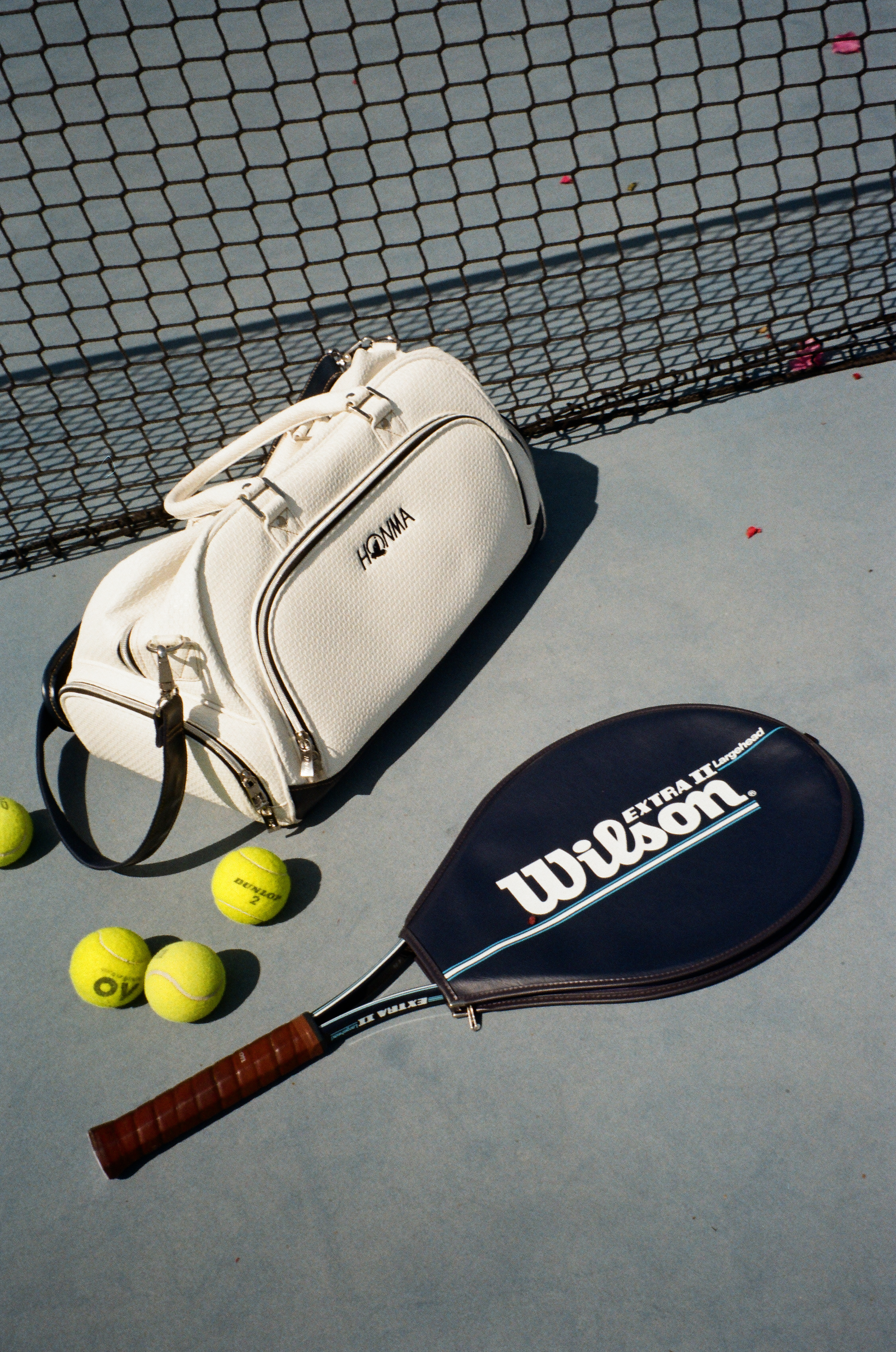 Equipo de tenis: bolsa Honma blanca, raqueta Wilson azul y pelotas en cancha.