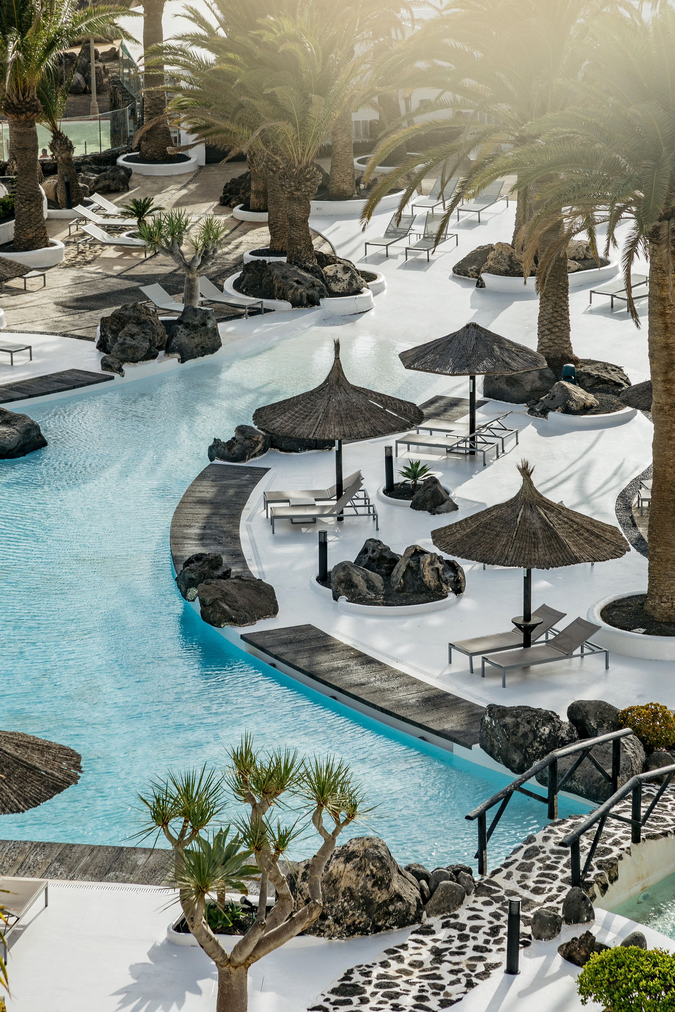 Tropical resort pool, white decks, palm trees, loungers, thatched umbrellas.