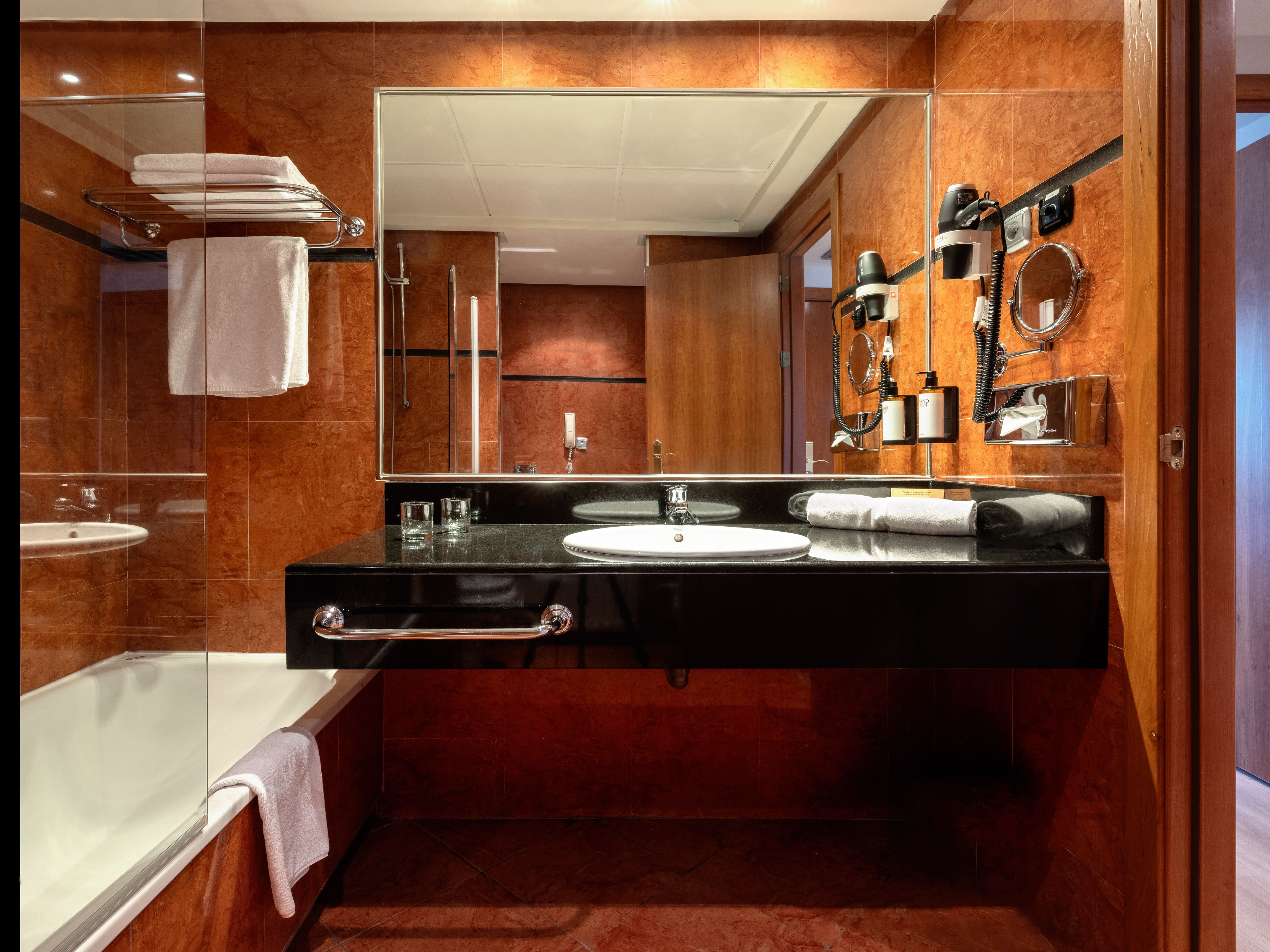Hotel bathroom featuring red tiles, black vanity, white sink, mirror, and bathtub.