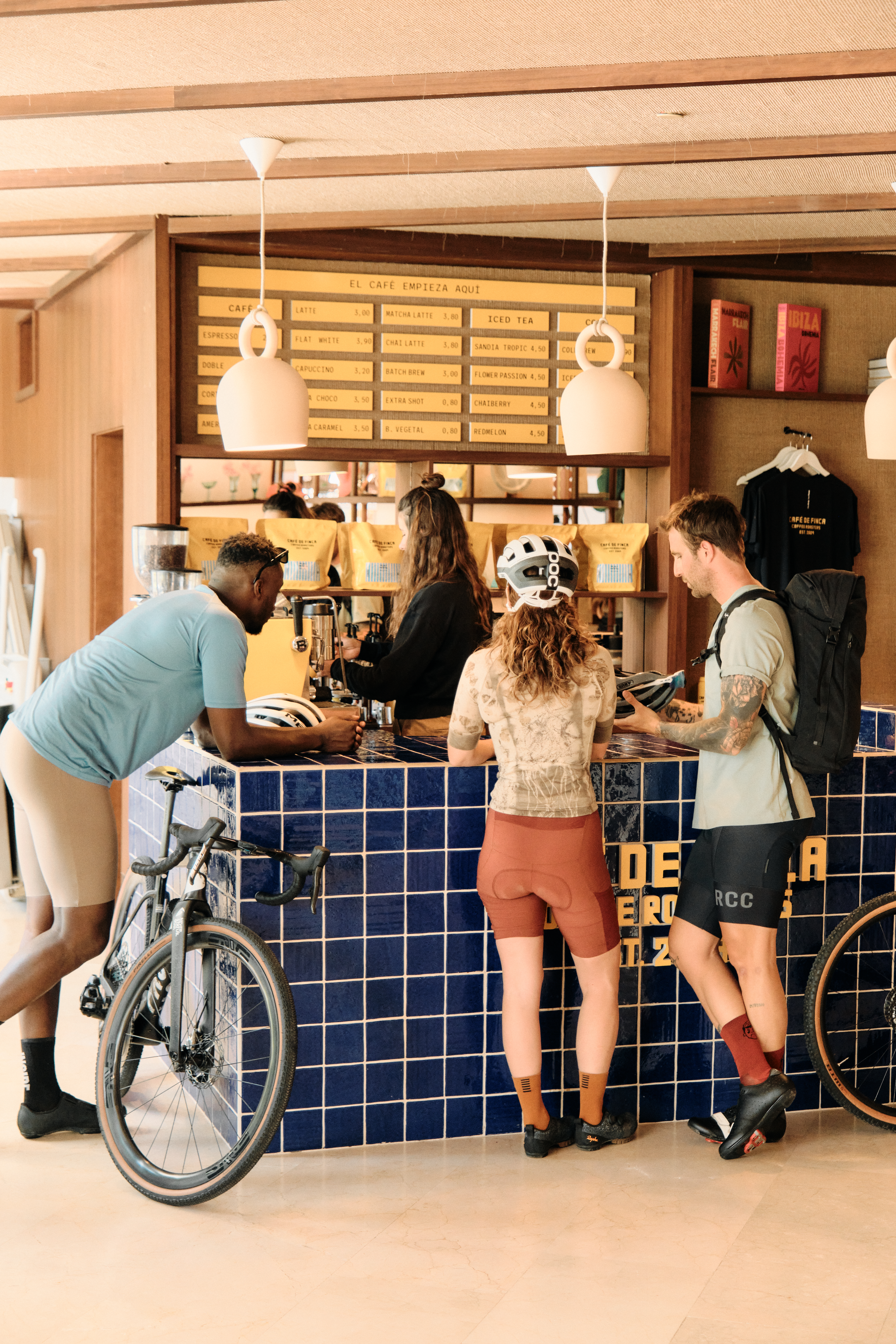 Cyclists in cycling gear ordering coffee at a cafe counter with bikes nearby.