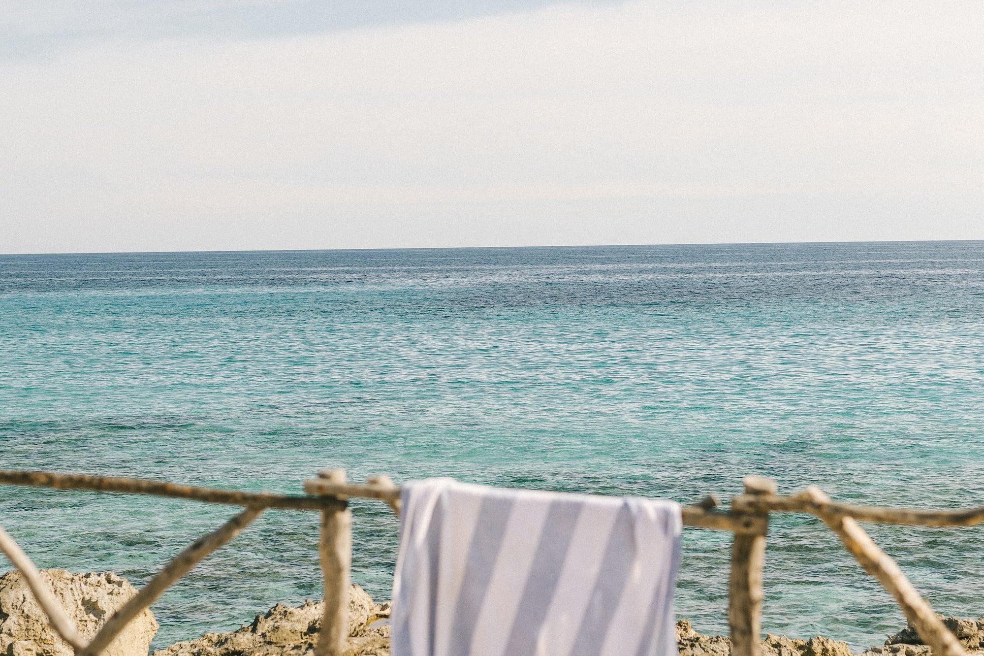 Toalla rayada en valla de madera con mar turquesa y costa rocosa bajo cielo azul.