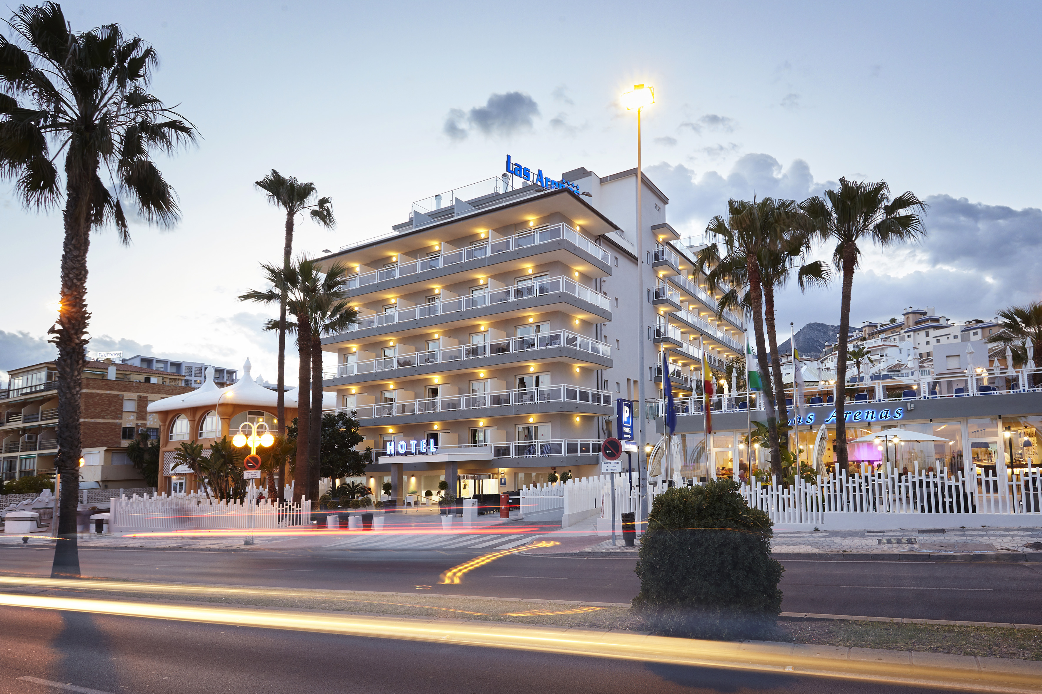 Coastal Las Arenas hotel illuminated at dusk with palm trees and traffic light trails.