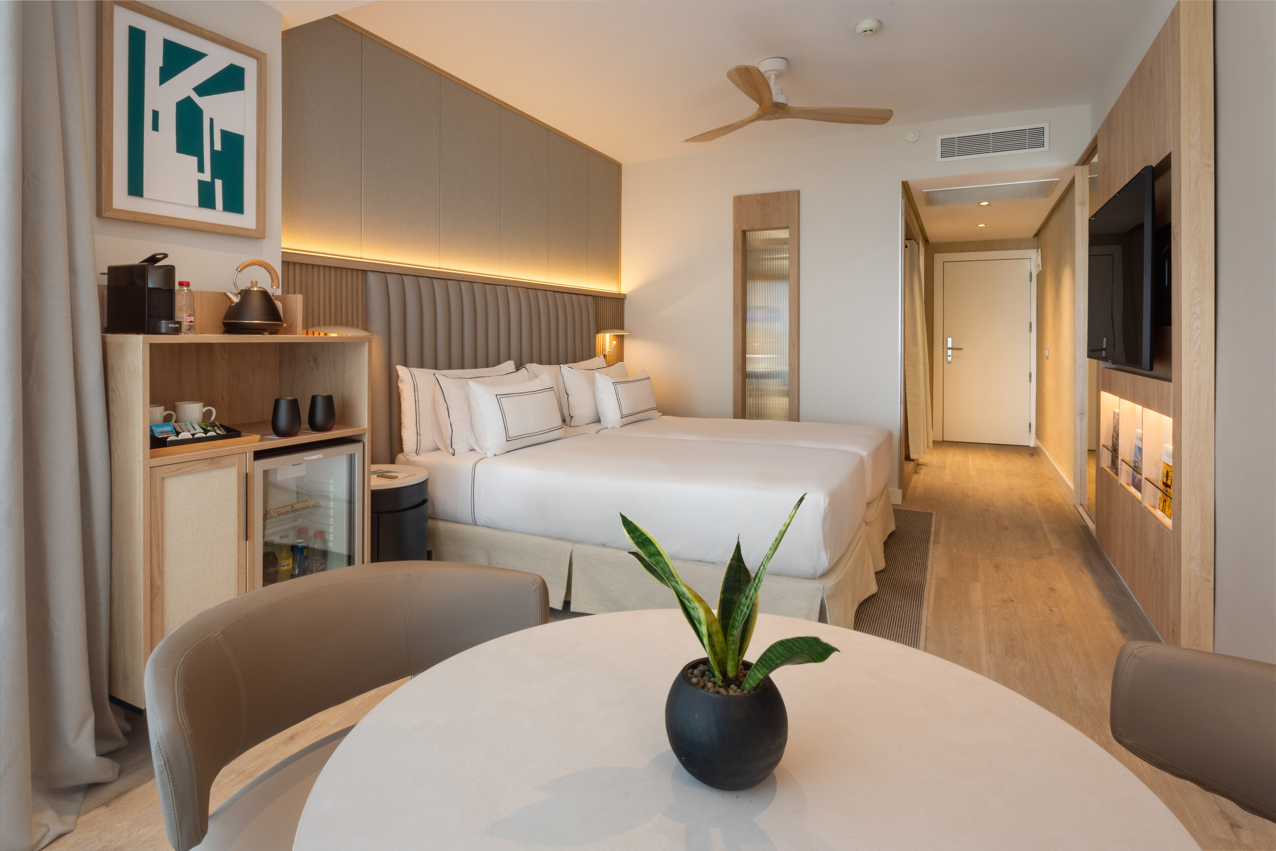 Modern hotel room with twin beds, minibar, ceiling fan, and contemporary decor.