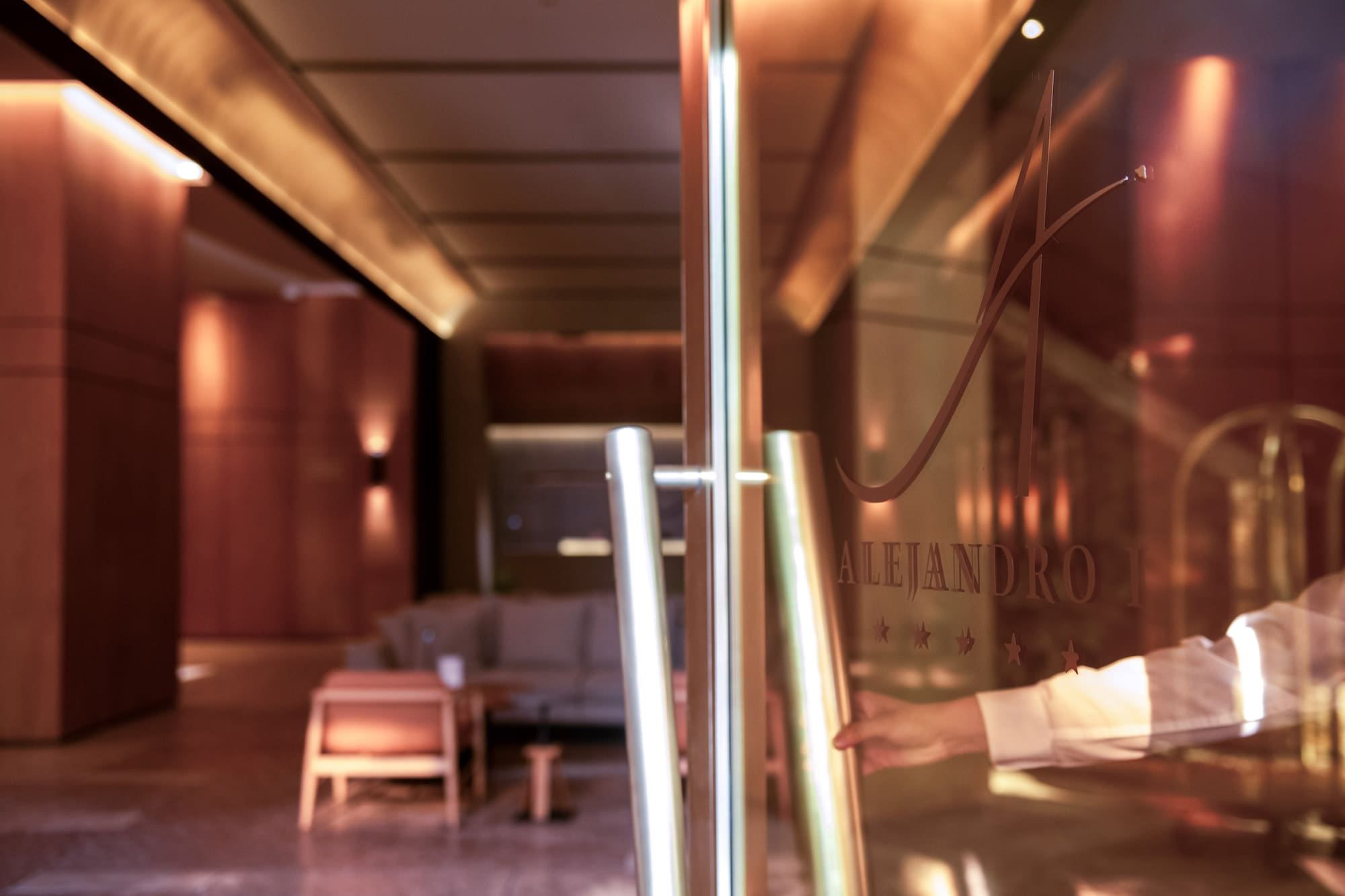 Hand opens modern glass hotel door to lobby, Alejandro logo.