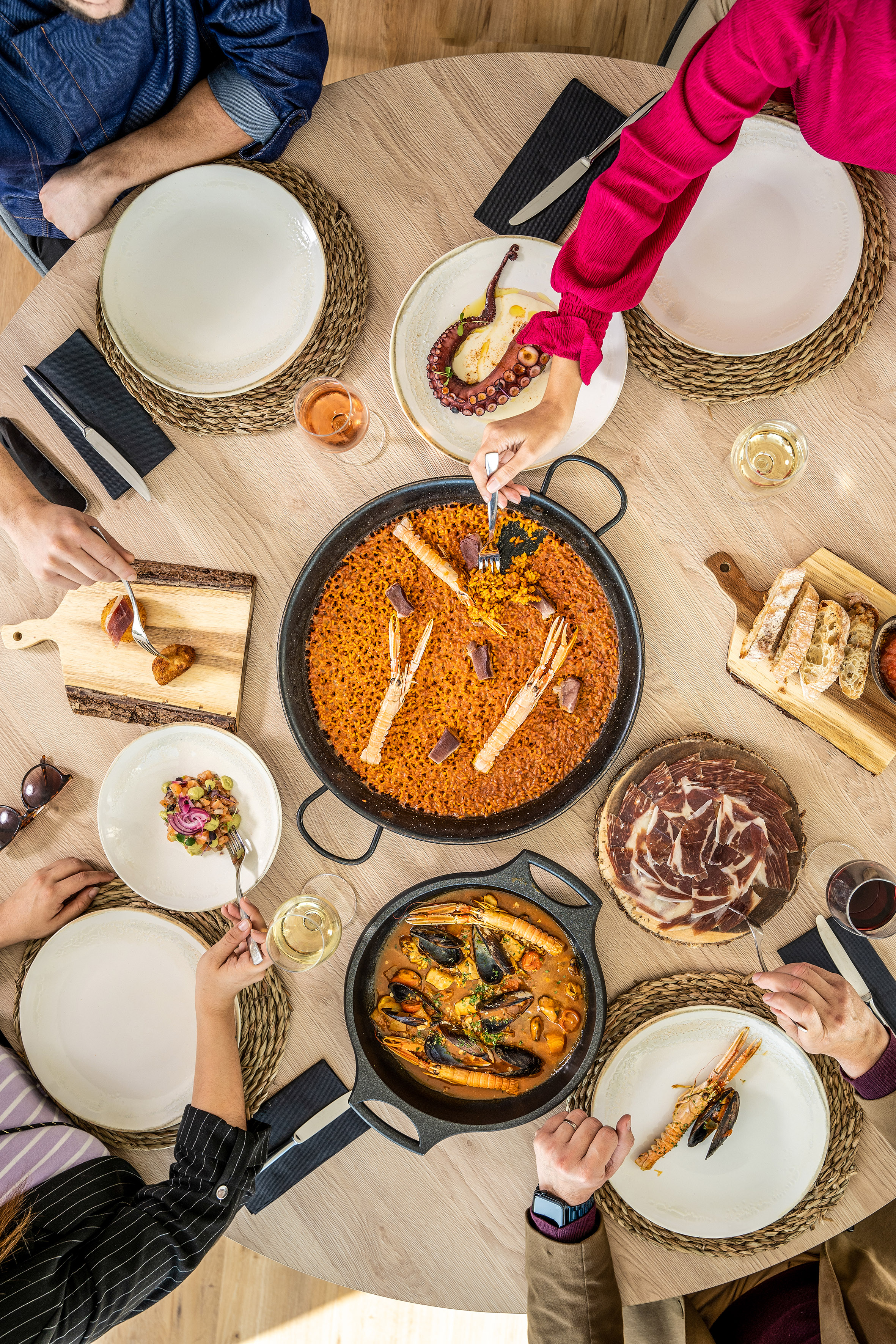 Paella, seafood, octopus, and ham on a wooden table with people dining.