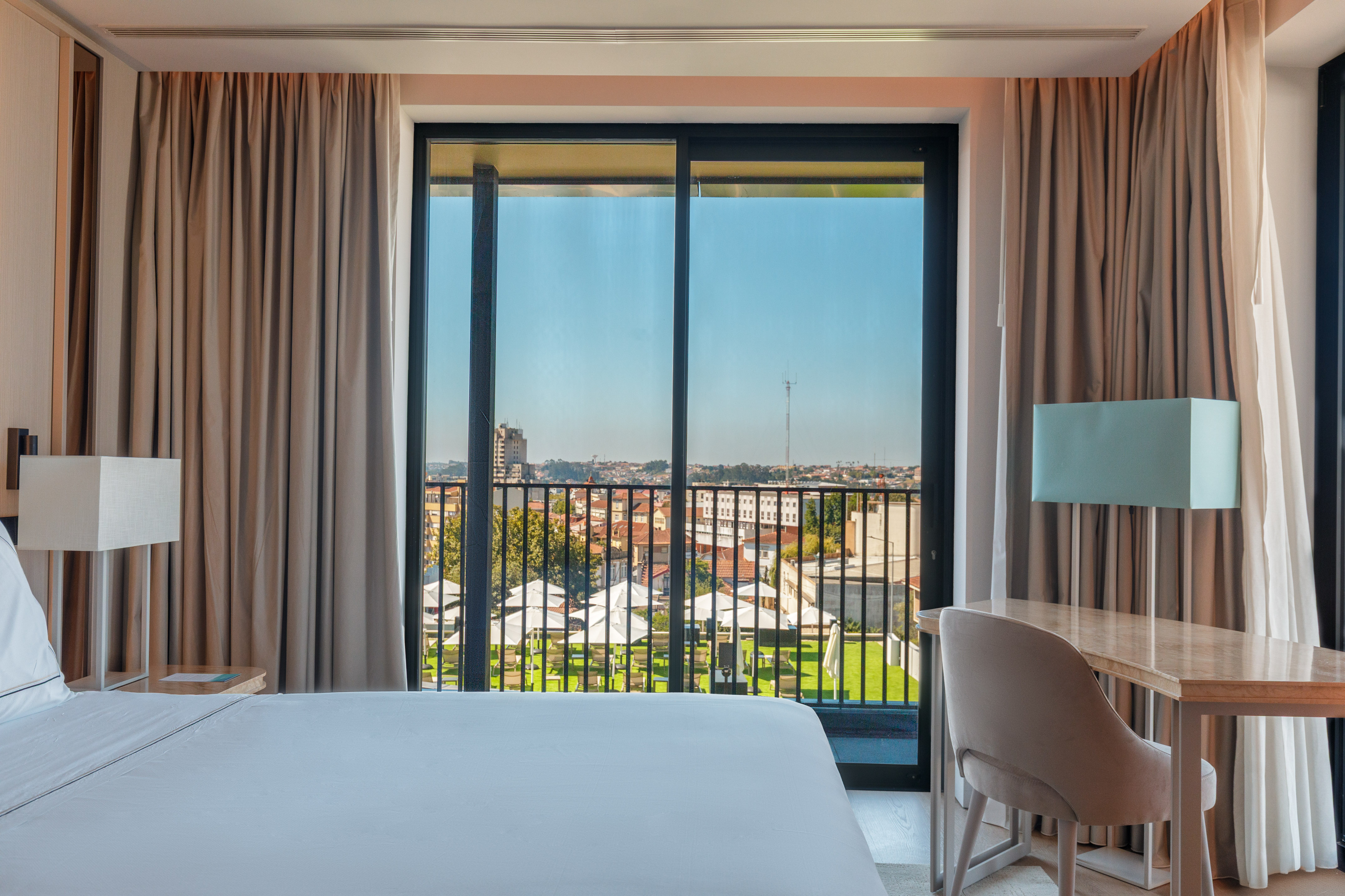 Modern hotel room with balcony city view, white bed, and workspace.