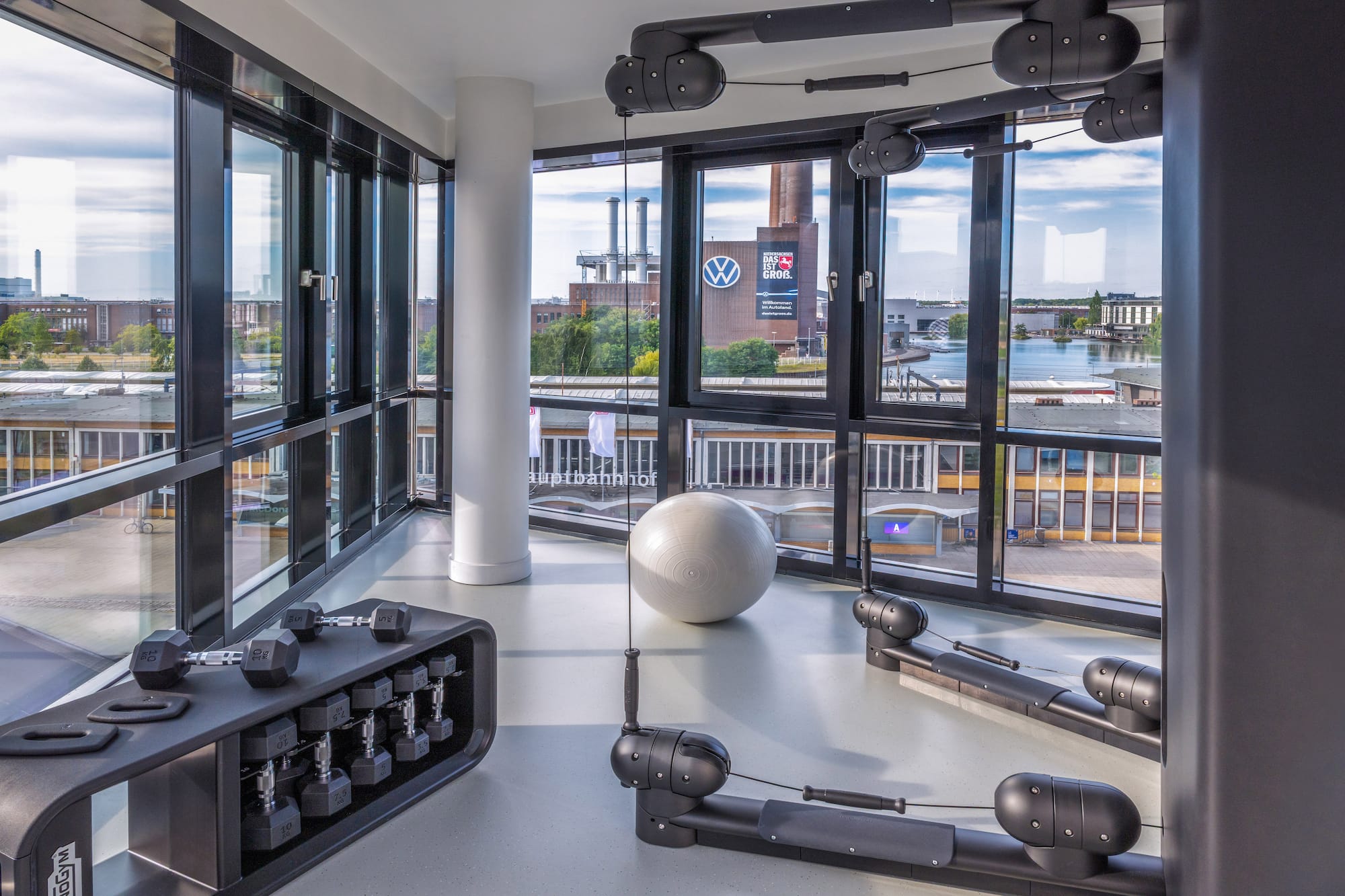 Modern gym with dumbbells, stability ball, and city view of Volkswagen plant.