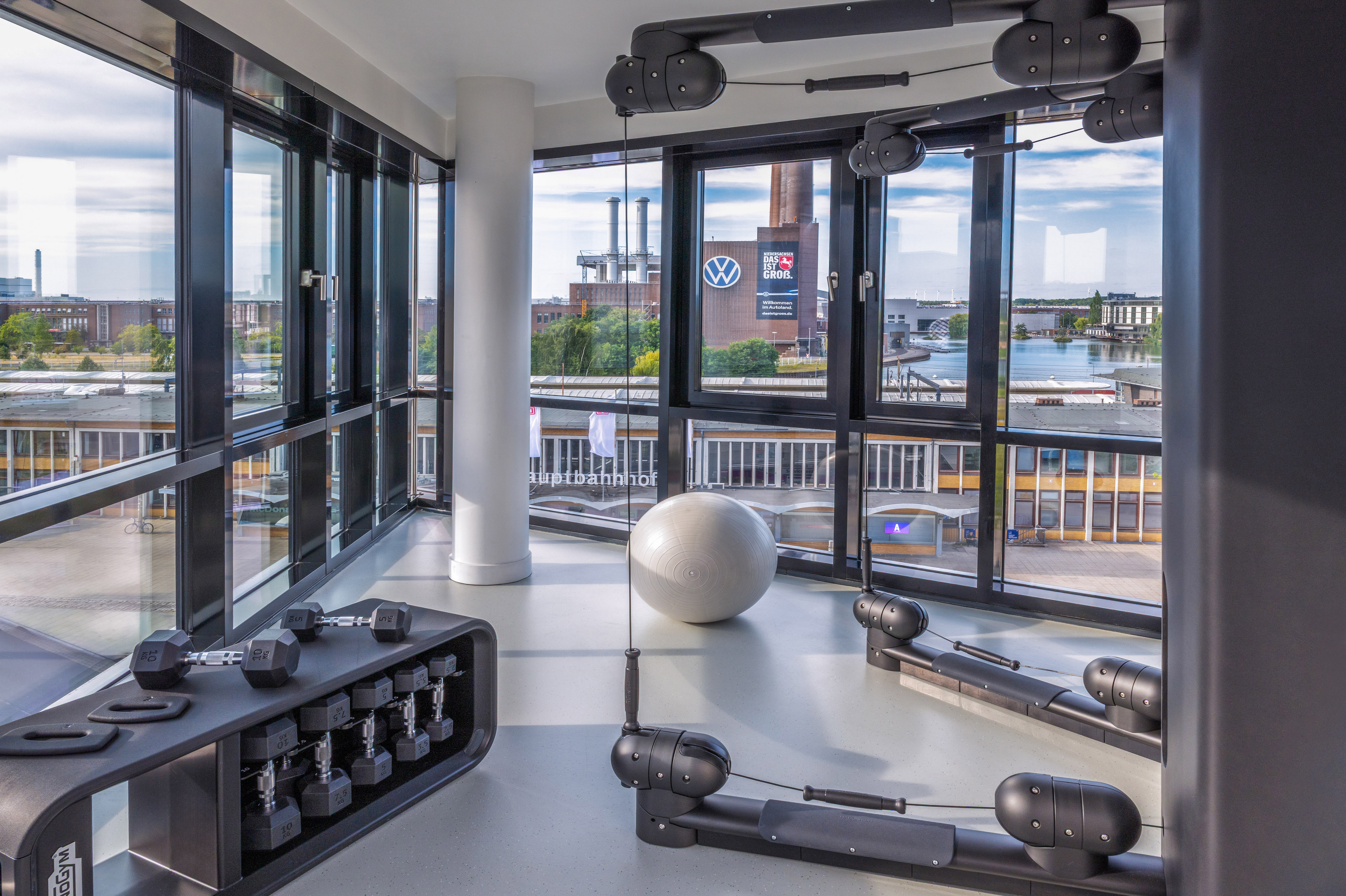 Modern gym with dumbbells, stability ball, and city view of Volkswagen plant.