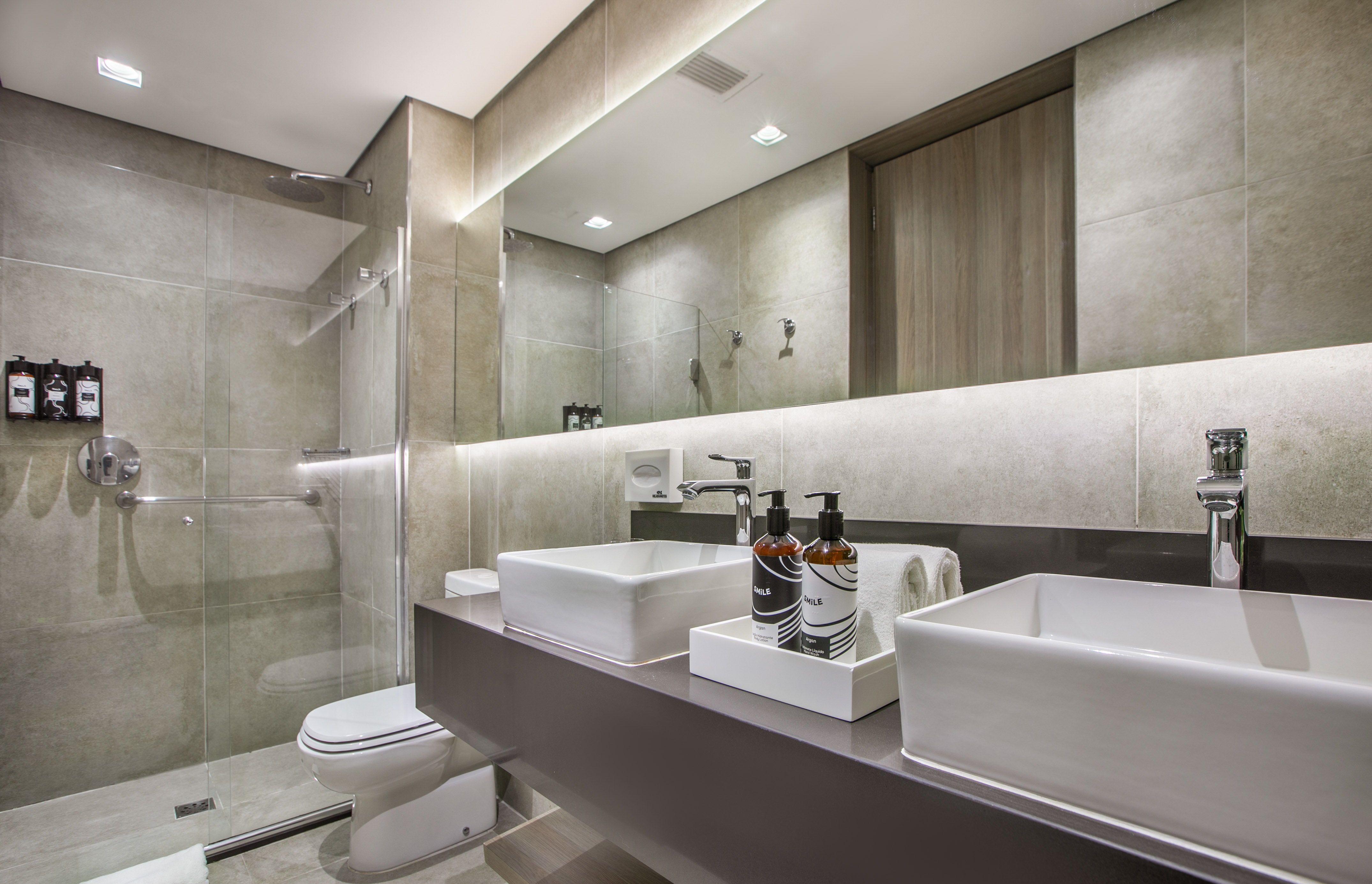Modern hotel bathroom with dual vessel sinks, walk-in shower, backlit mirror.