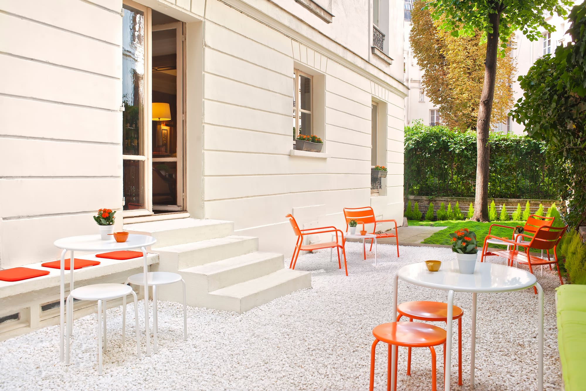 Modern outdoor patio with white gravel, orange chairs, white tables, and green garden.