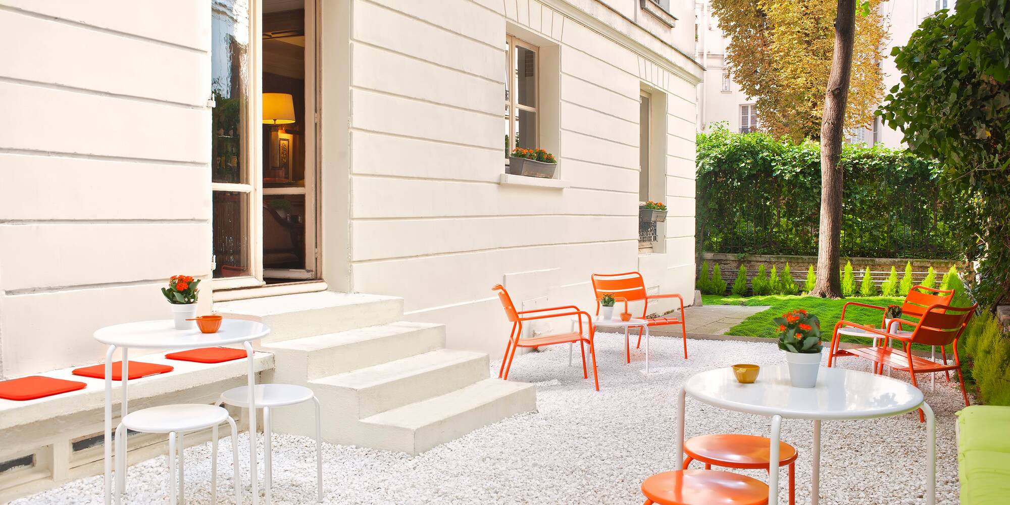 Modern outdoor patio with white gravel, orange chairs, white tables, and green garden.