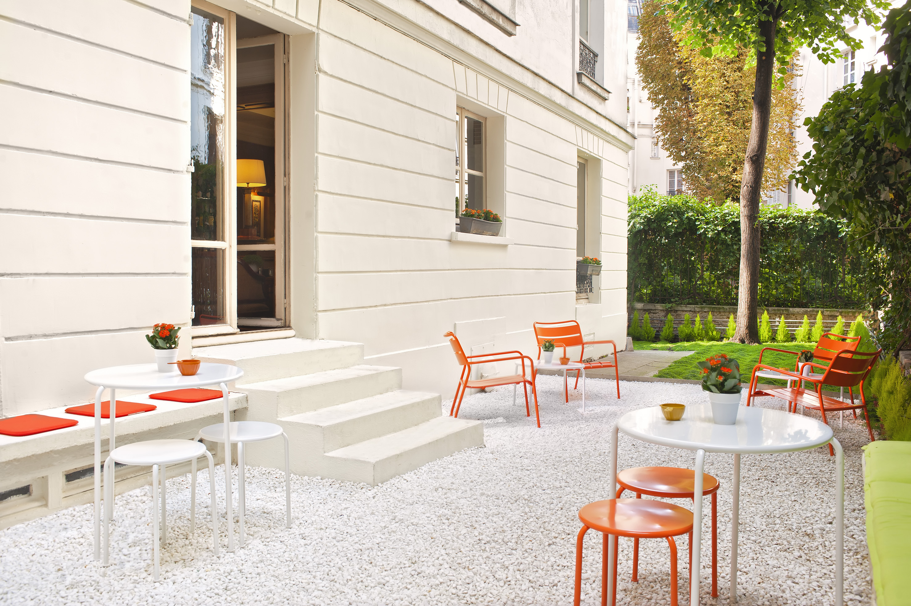 Modern outdoor patio with white gravel, orange chairs, white tables, and green garden.