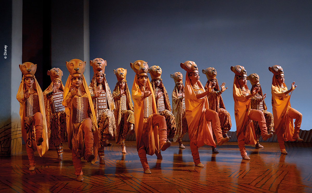 Lion King musical performers in ornate lion mask costumes on stage.