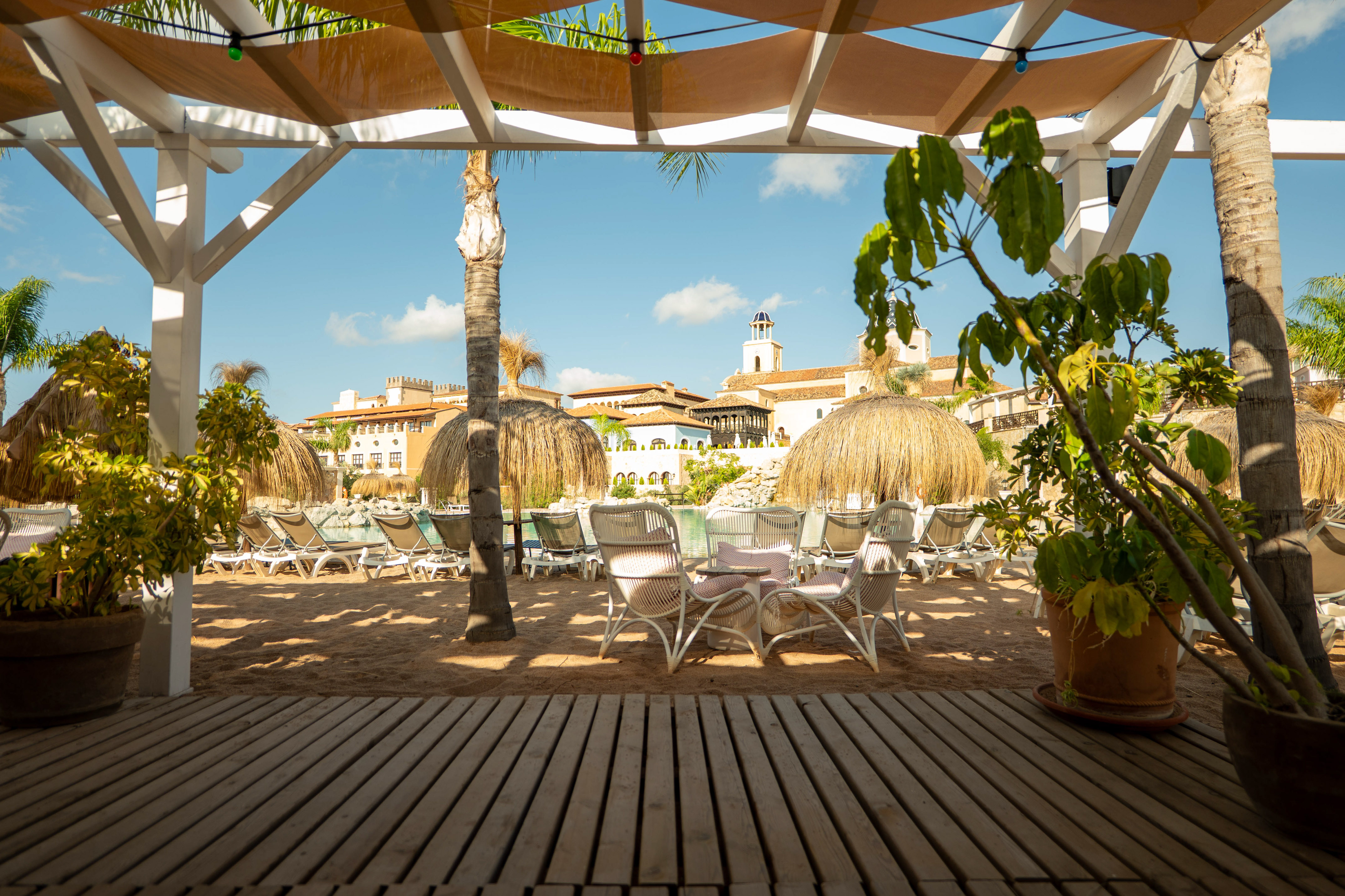 Shaded resort beach area with sand, pool, thatched umbrellas, and hotel buildings.