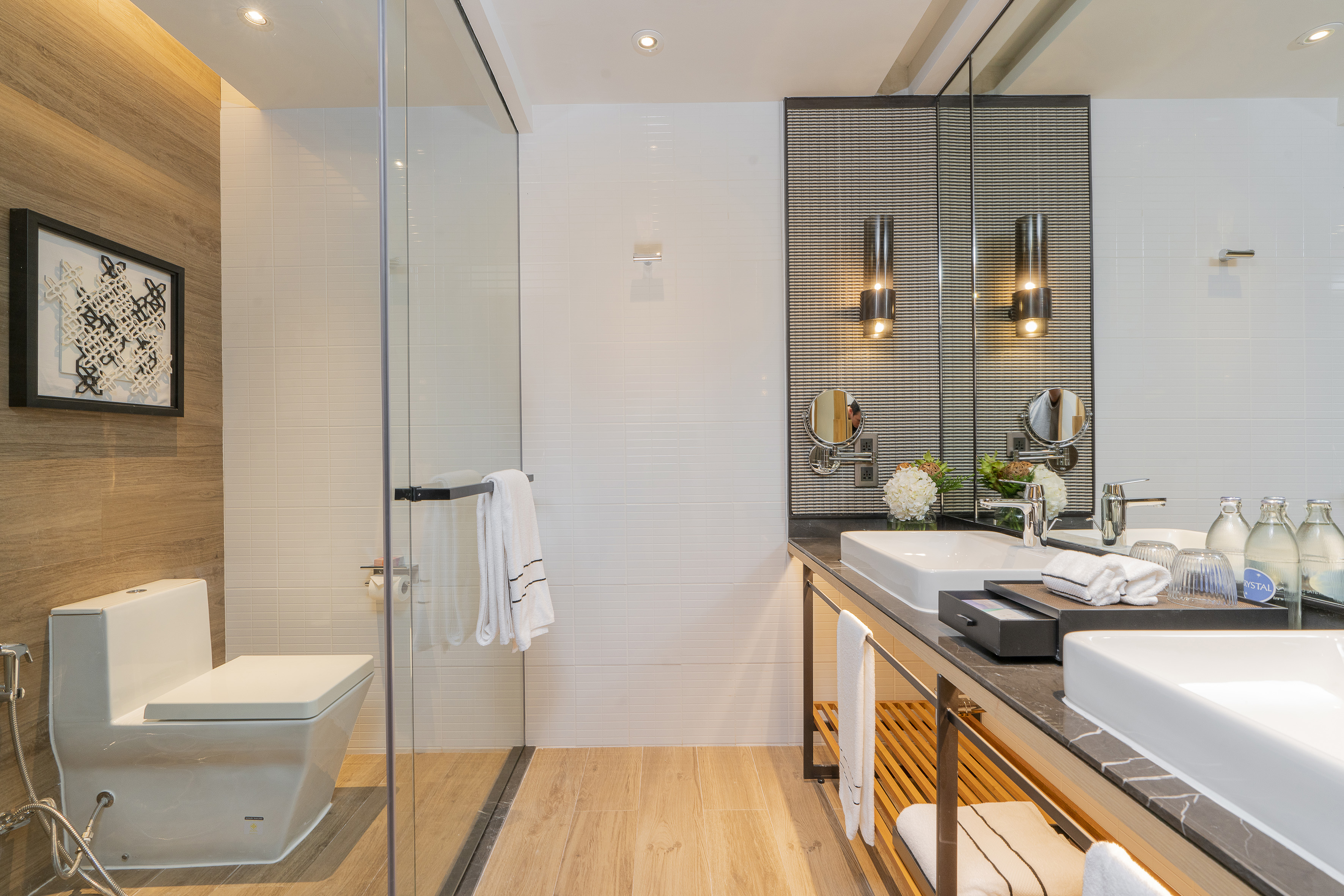 Modern hotel bathroom with wood accents, glass shower, toilet, and double vanity.