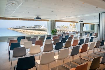 Sala conferencias moderna, vista al mar con muelle, sillas, proyector.