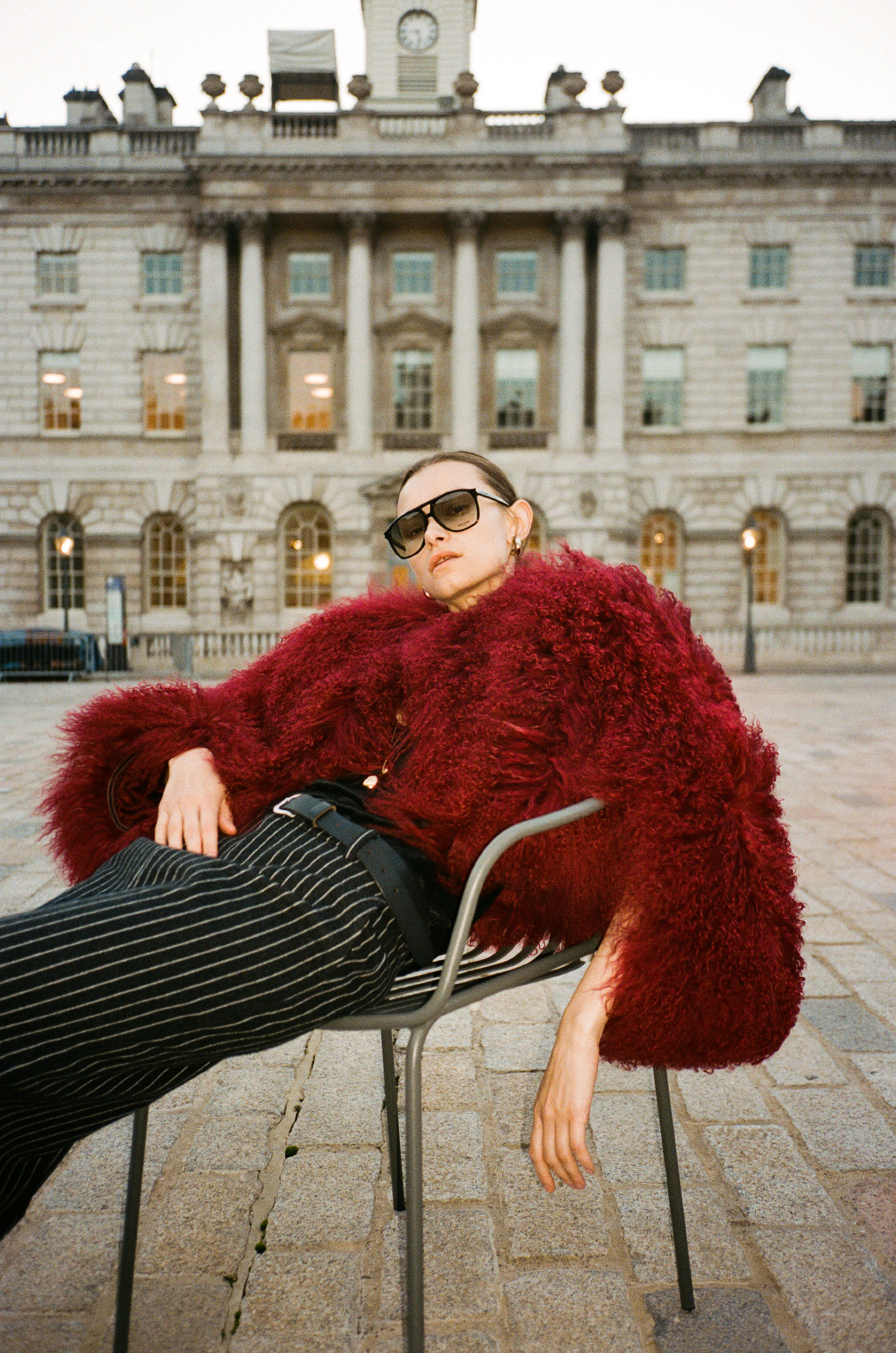 Woman in red fluffy coat and sunglasses, by a grand stone building.