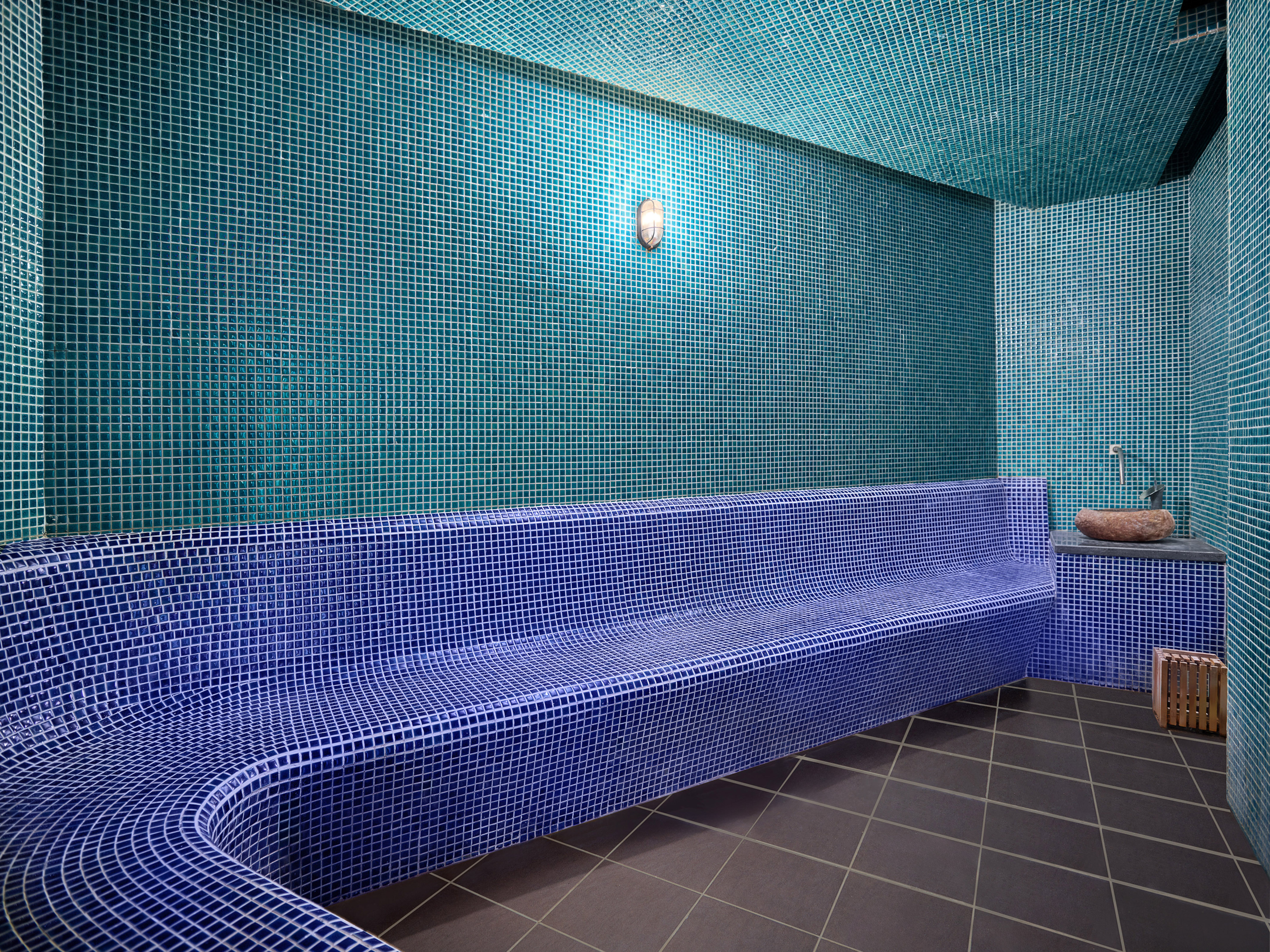 Blue mosaic tiled steam room with teal walls, curved bench, and stone sink.