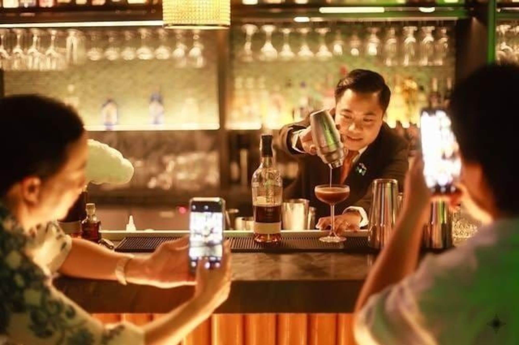Mixologist pours cocktail, patrons photograph with smartphones in a dim bar.