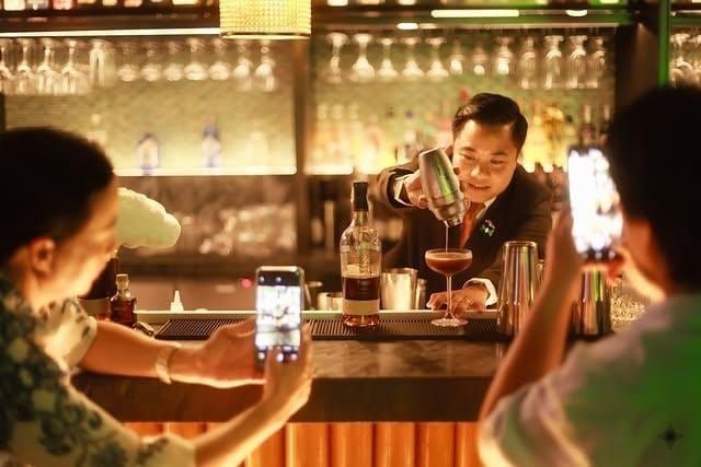 Mixologist pours cocktail, patrons photograph with smartphones in a dim bar.