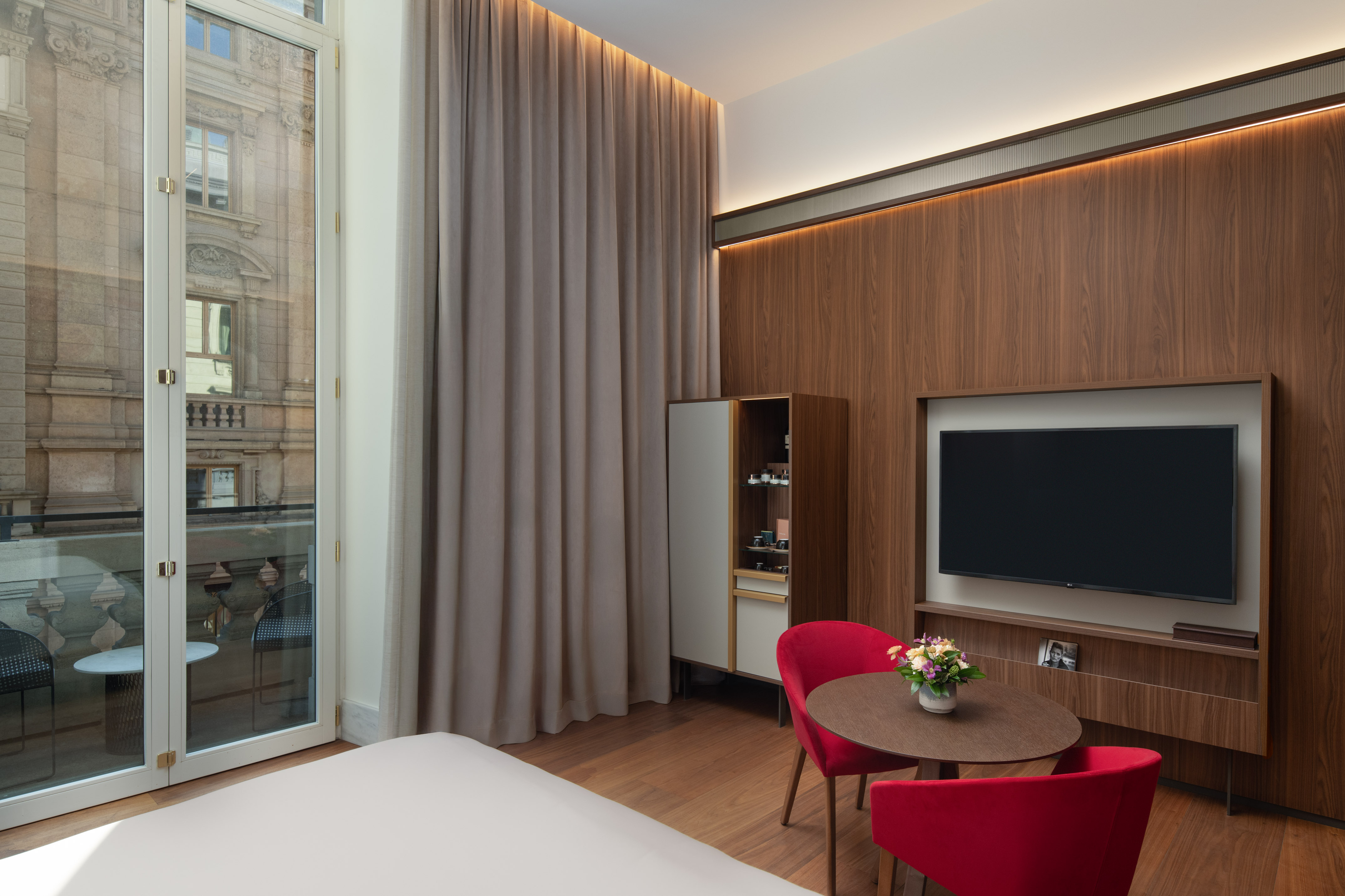 Modern hotel room with balcony city view, wood paneling, TV, and red chairs.