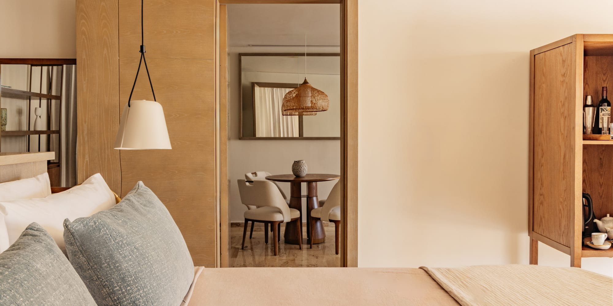 Modern hotel bedroom, view to living area with wood table, wicker light.