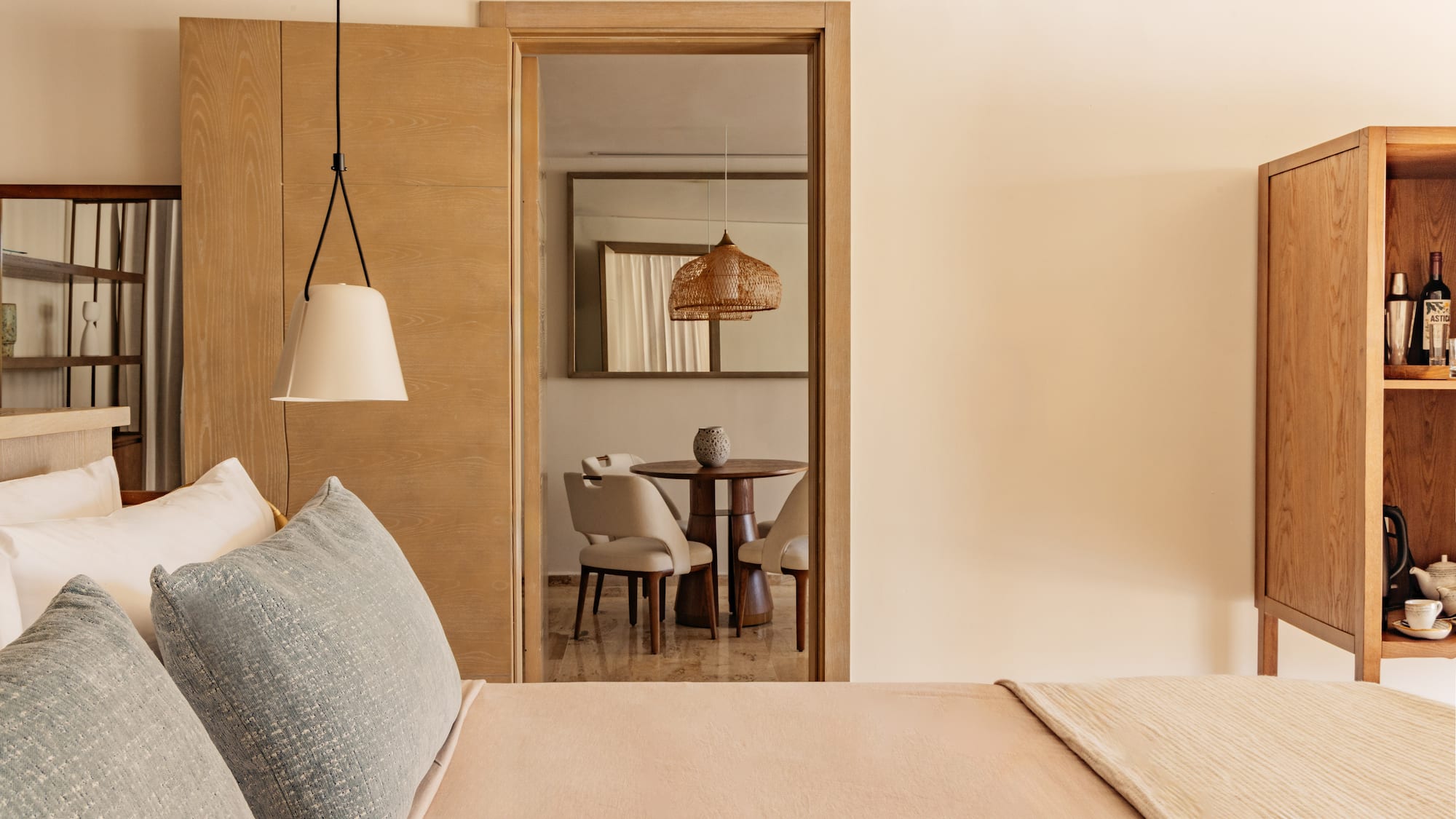 Modern hotel bedroom, view to living area with wood table, wicker light.