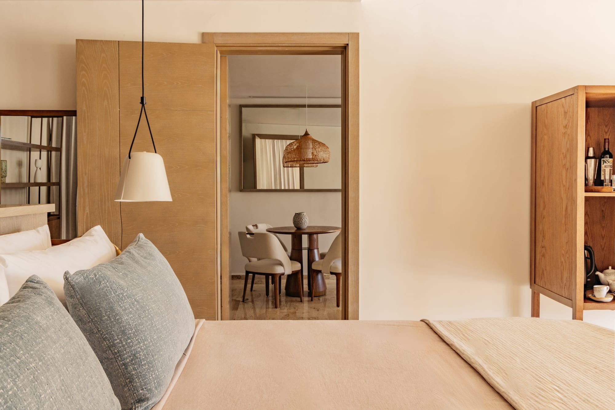 Modern hotel bedroom, view to living area with wood table, wicker light.