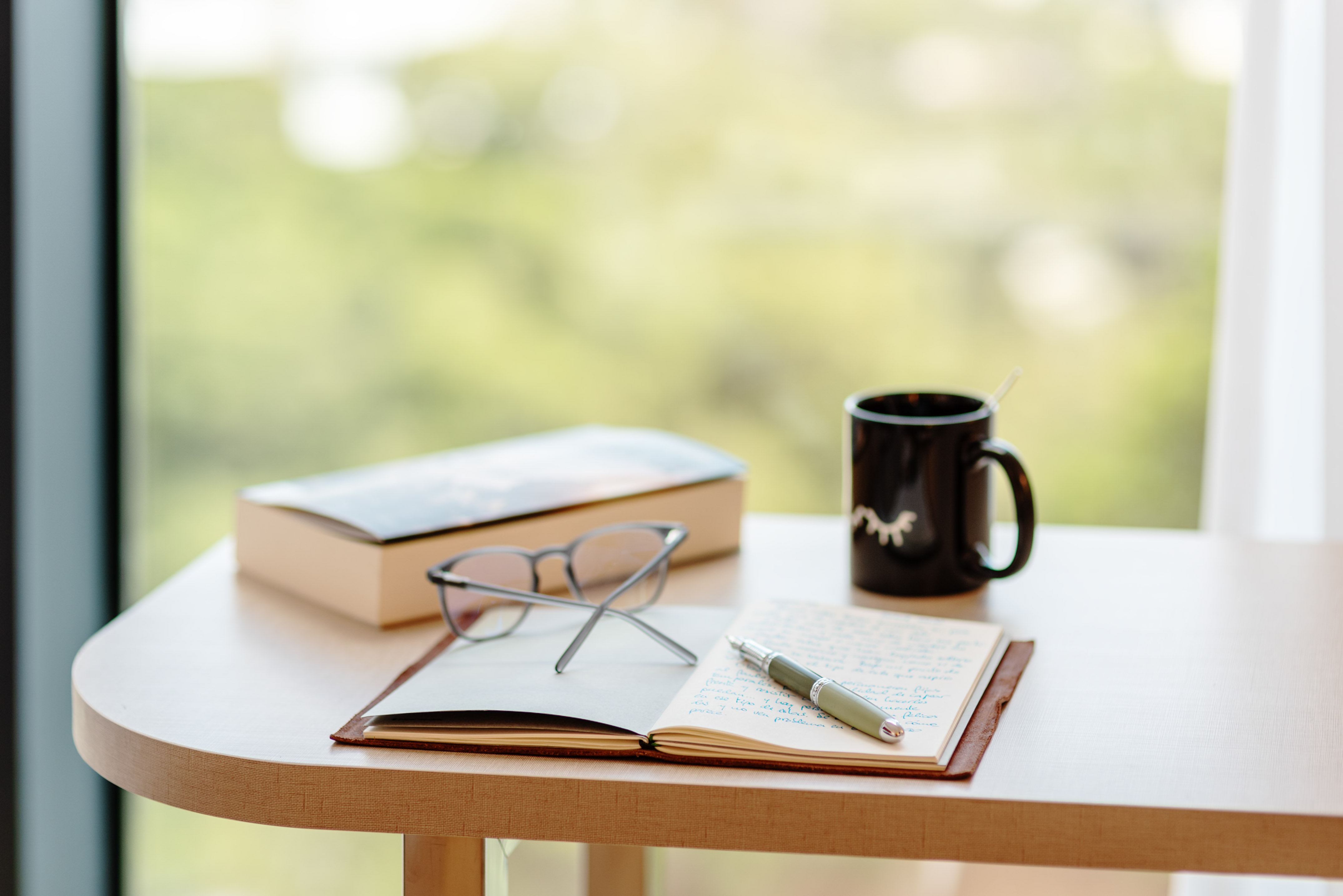 Workstation: notebook, pen, eyeglasses, book, coffee mug by window.