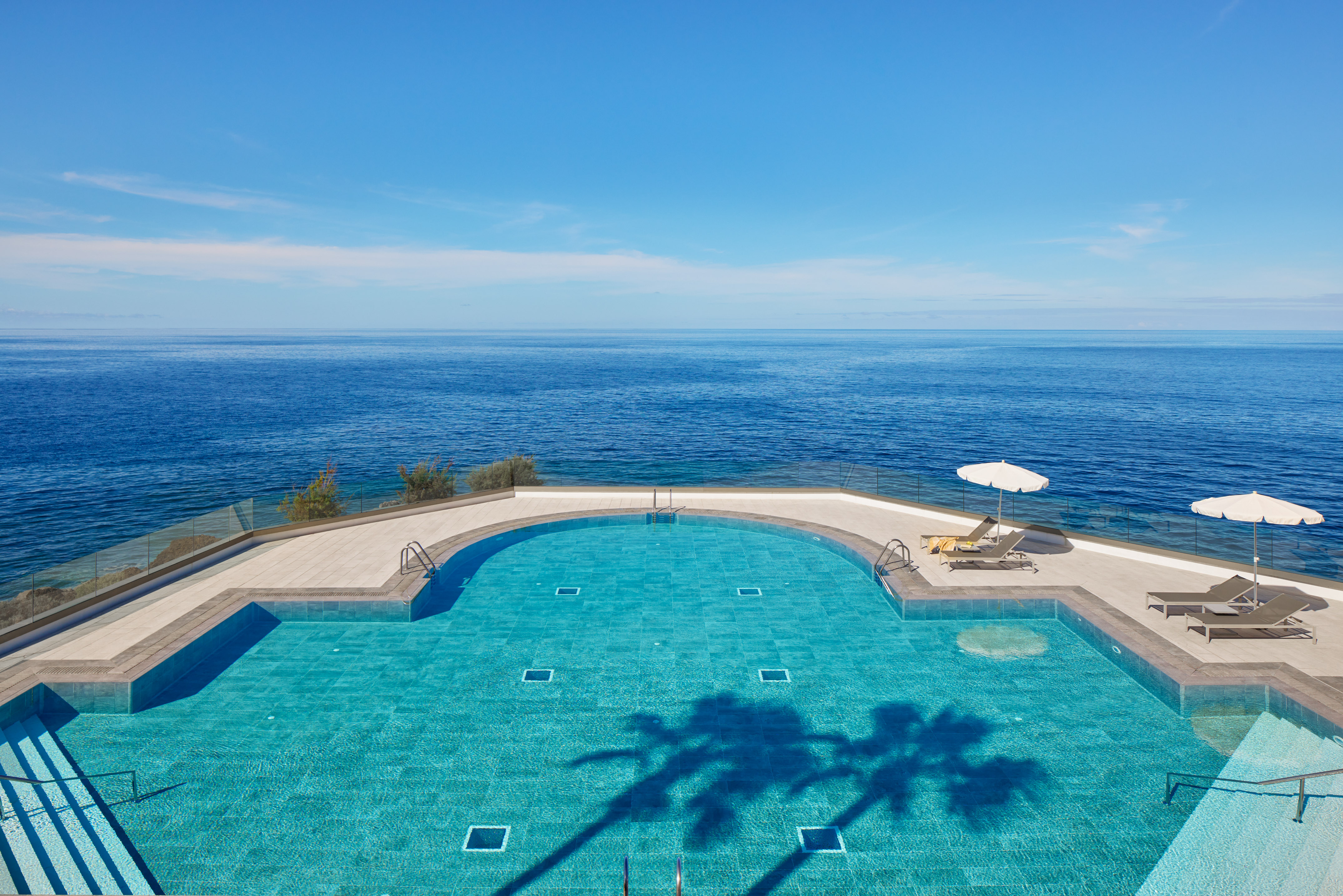 Infinity pool overlooking blue ocean with sun loungers and palm shadow.