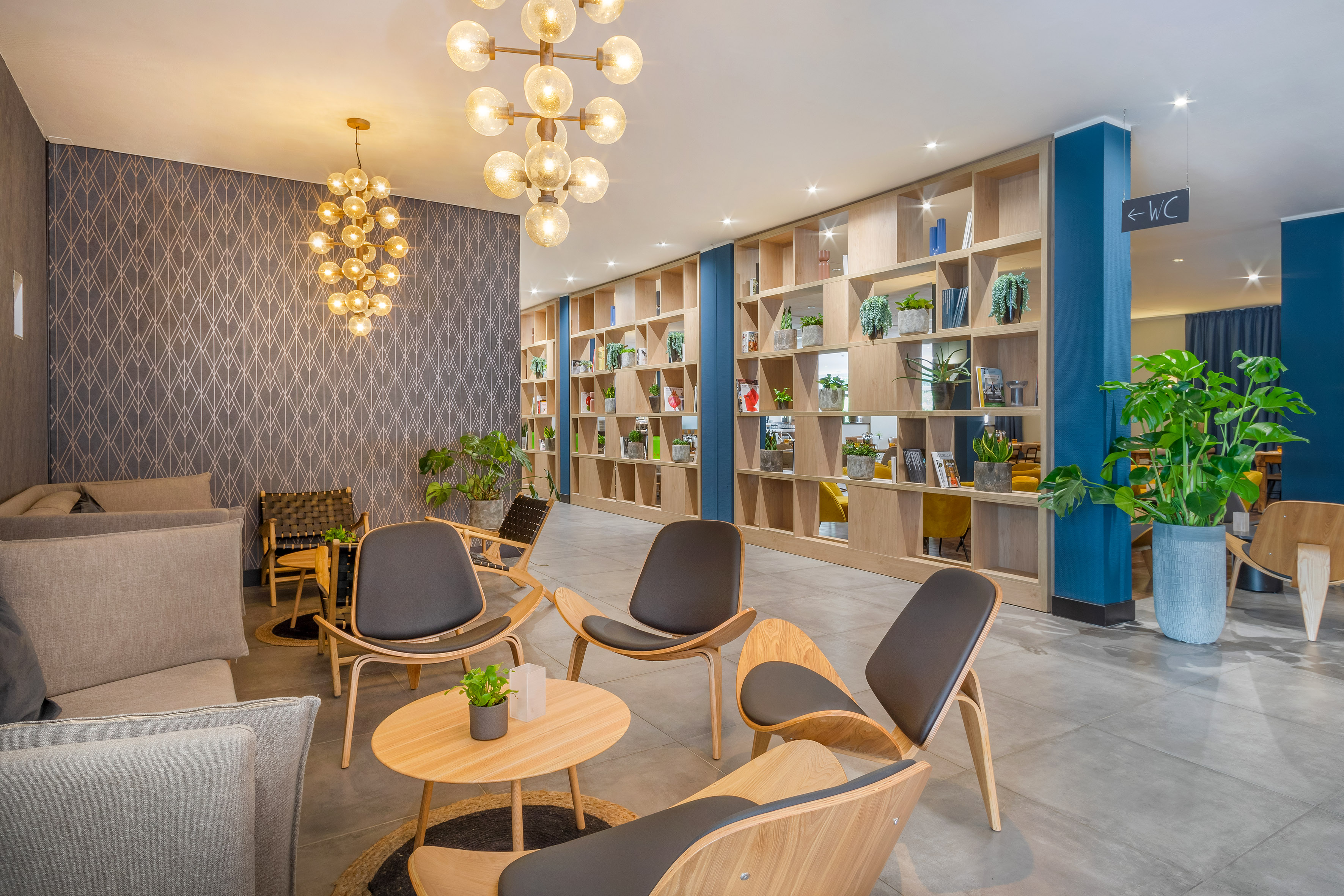 Contemporary hotel lounge with wooden chairs, plant shelving, and chandeliers.