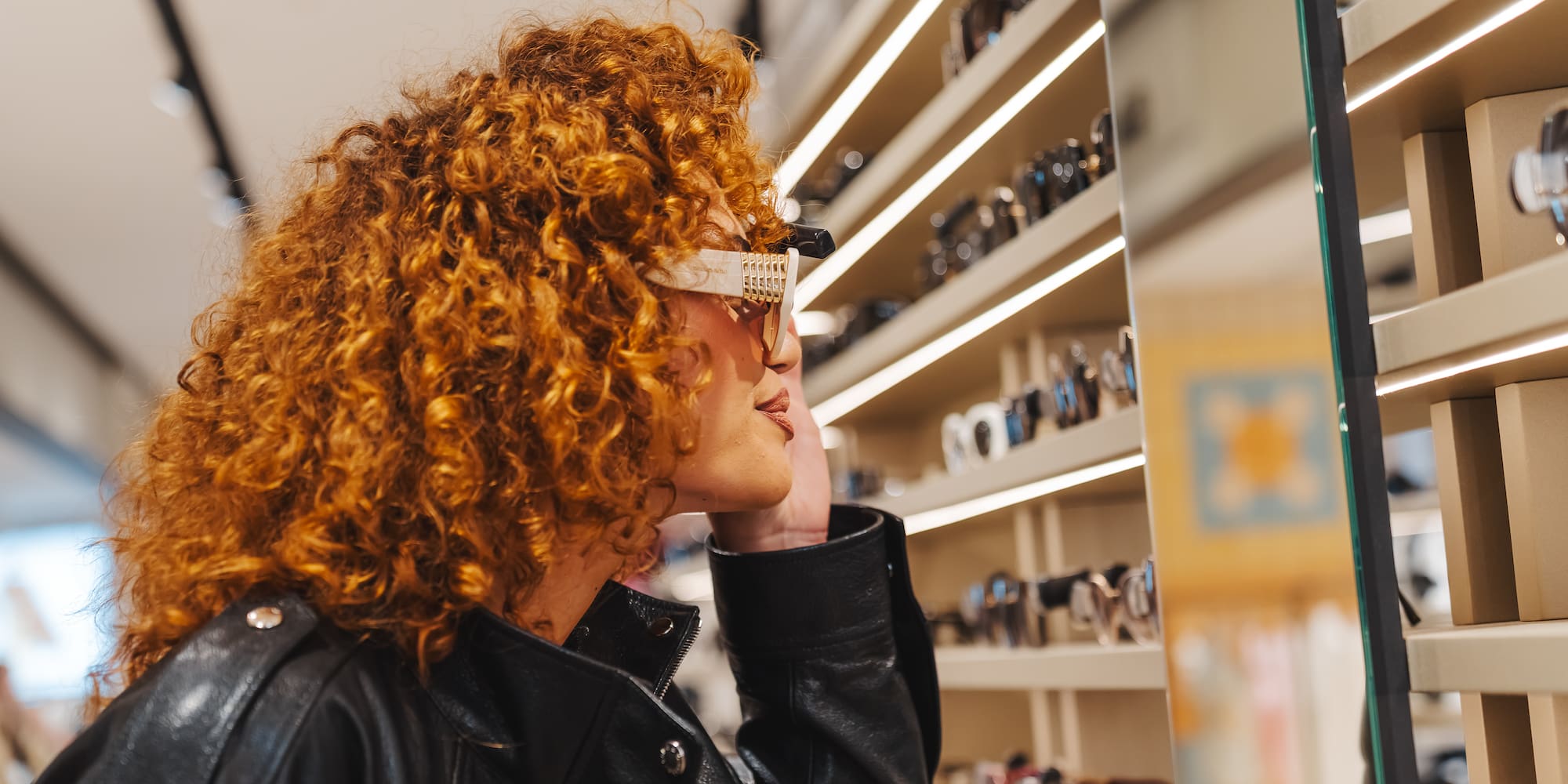 Curly red-haired woman trying on white sunglasses in an eyewear store.