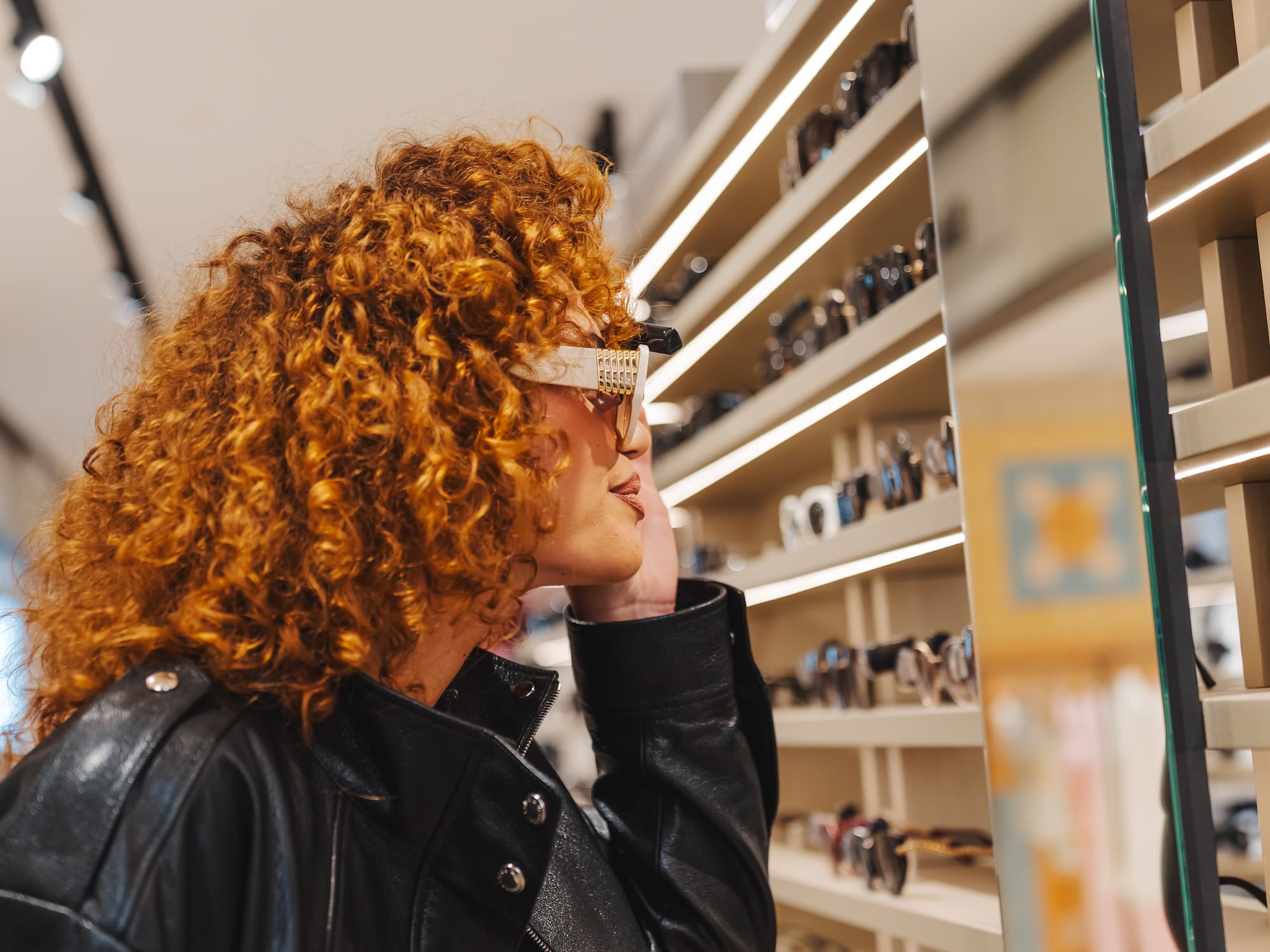 Curly red-haired woman trying on white sunglasses in an eyewear store.