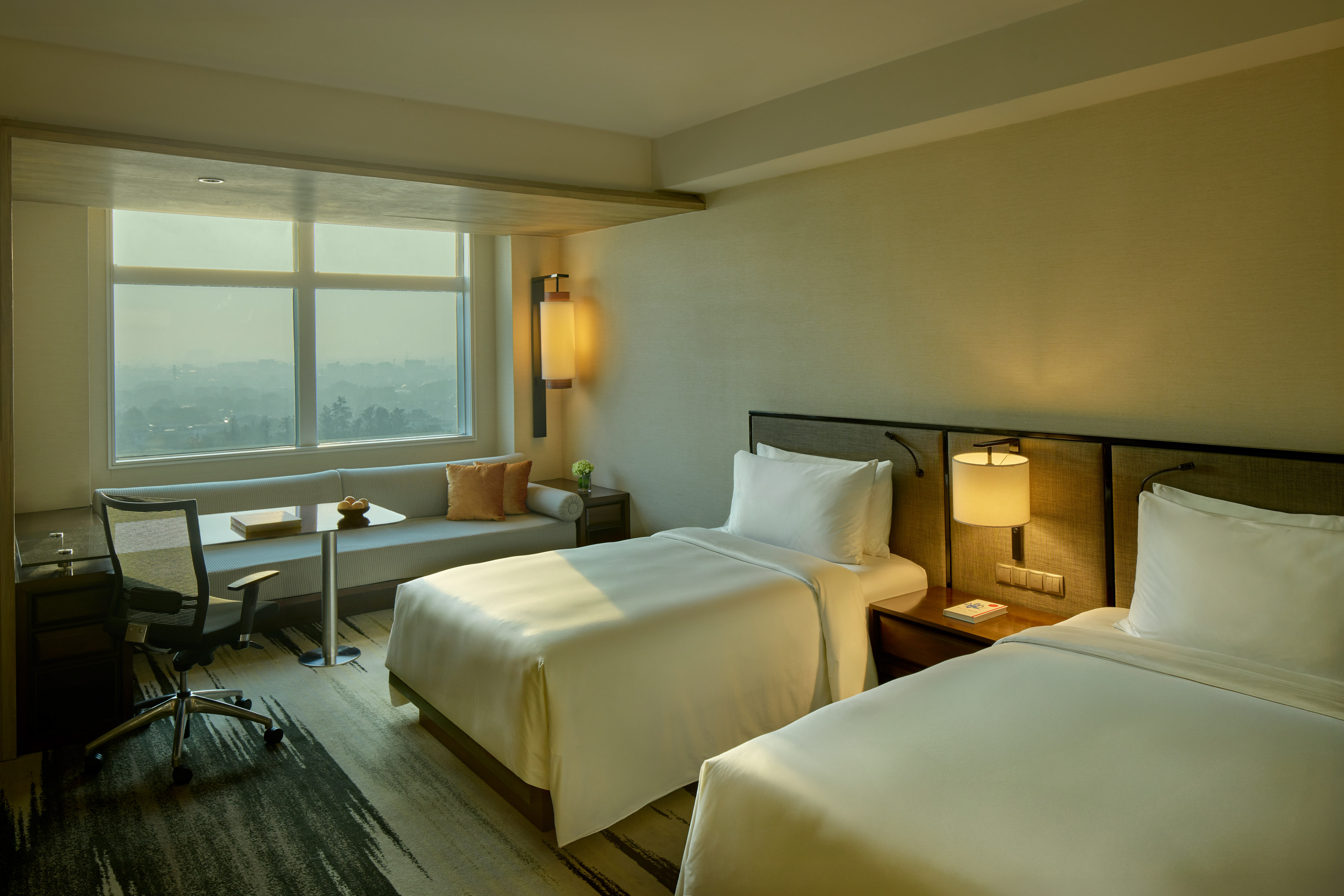 Modern hotel room with twin beds, large window city view, and desk.