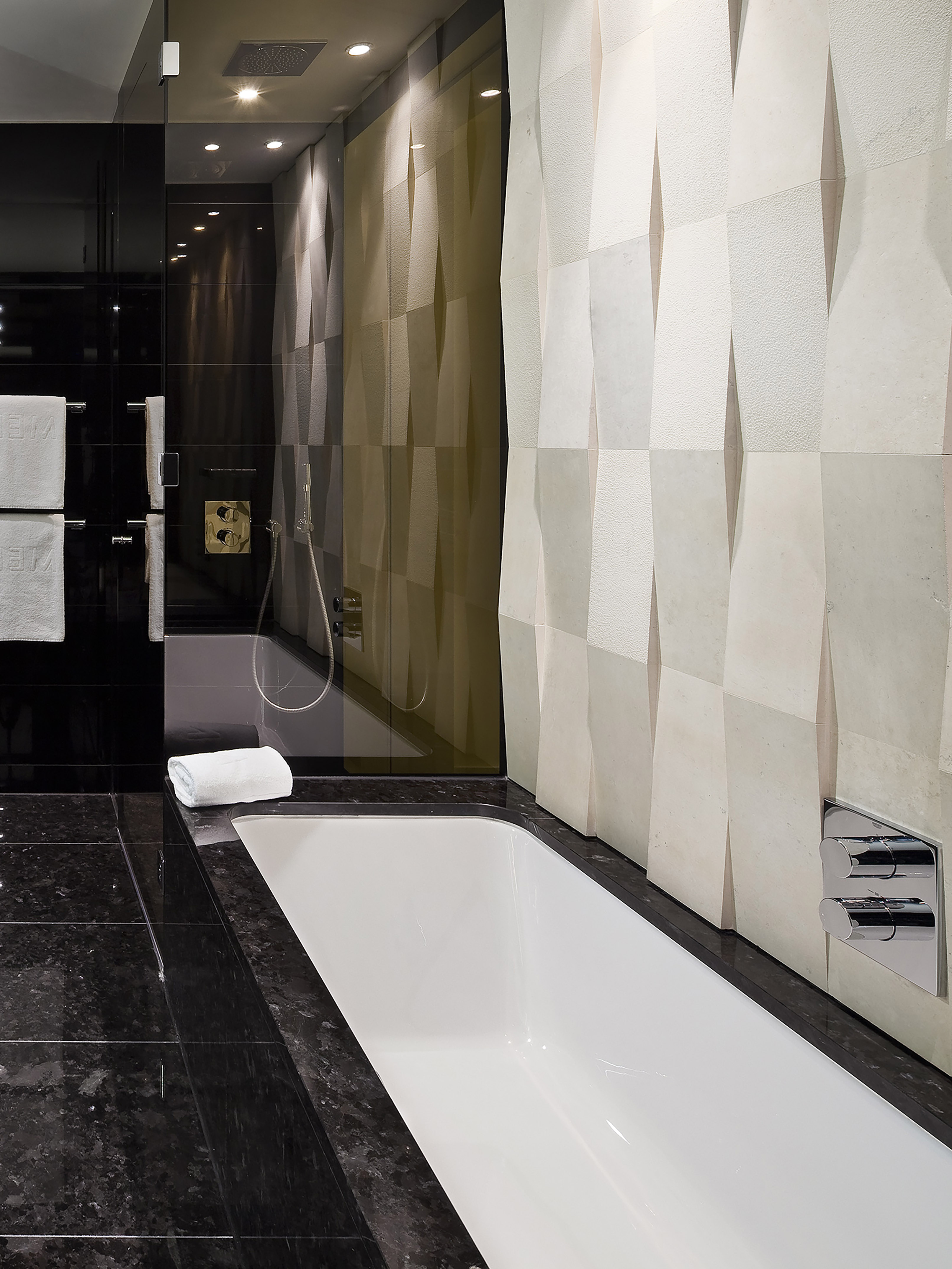 Modern bathroom with white bathtub, glass shower, black marble, and geometric wall tiles.