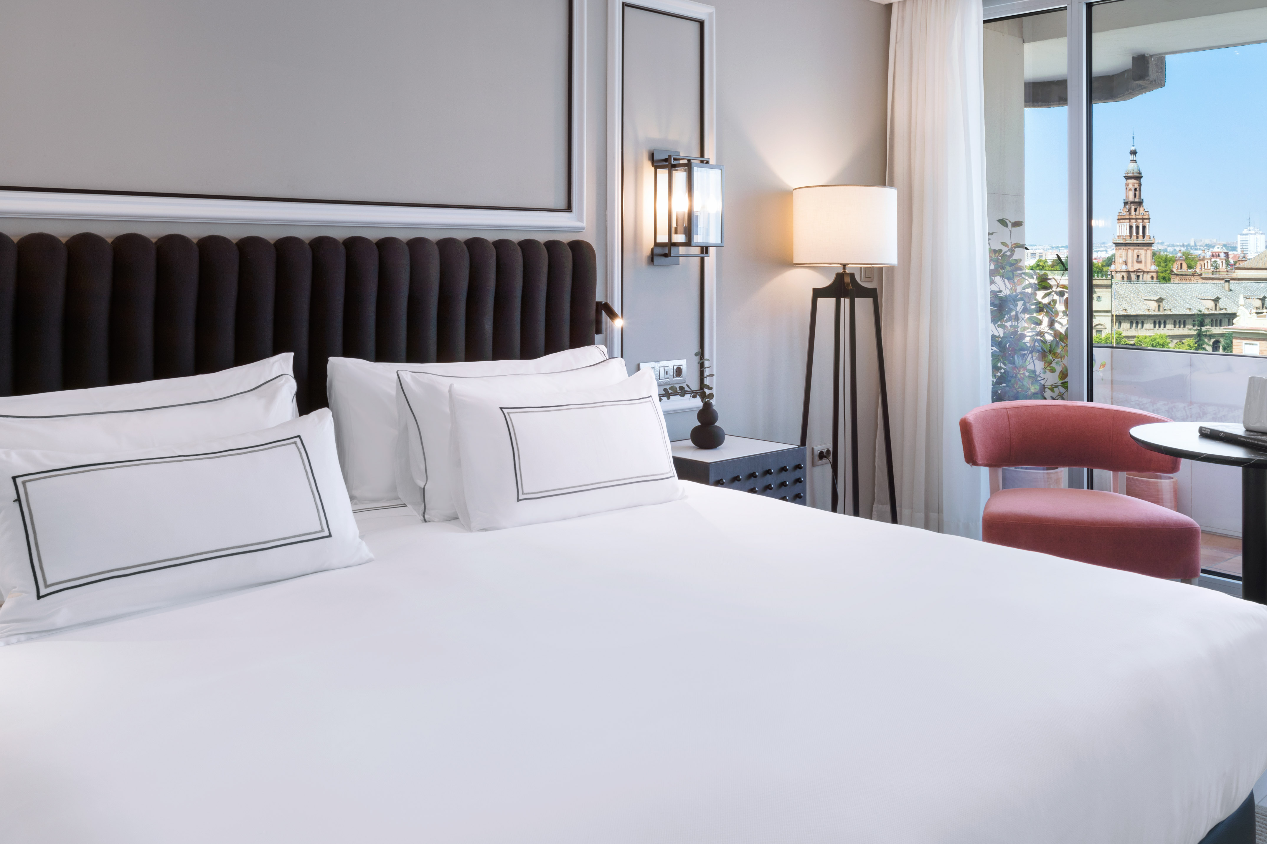 Modern hotel bedroom with white bed, dark headboard, and city view balcony.